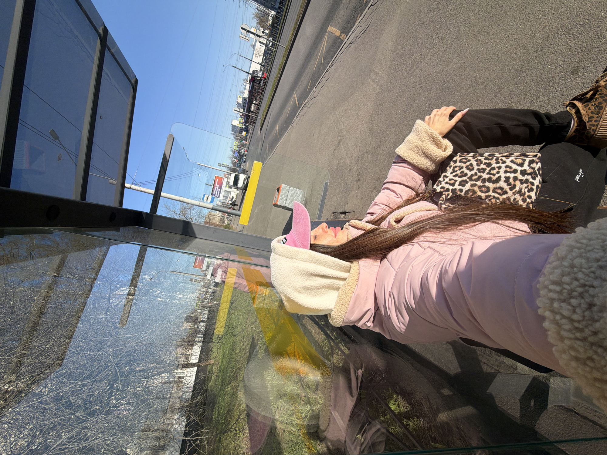 A person lies on a bench at a bus stop. The bus stop has glass sides, and the reflection of trees, grass, and buildings can be seen. Outside the bus stop, there is a road with cars, buildings, and power lines. The person is wearing a pink jacket with a beige sherpa lining, black pants, leopard print shoes, and a pink and white hooded hat. They also have a leopard print purse. The location is Saint Petersburg, Russia.
