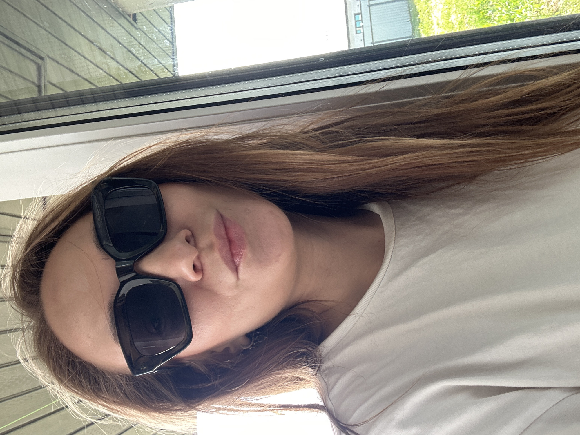 A woman with brown hair and sunglasses poses indoors near a window. The background shows a partial view of buildings and greenery through the glass. The subject is wearing a white shirt. There is no information in the image that would identify the location as Kharnavy, Russia.