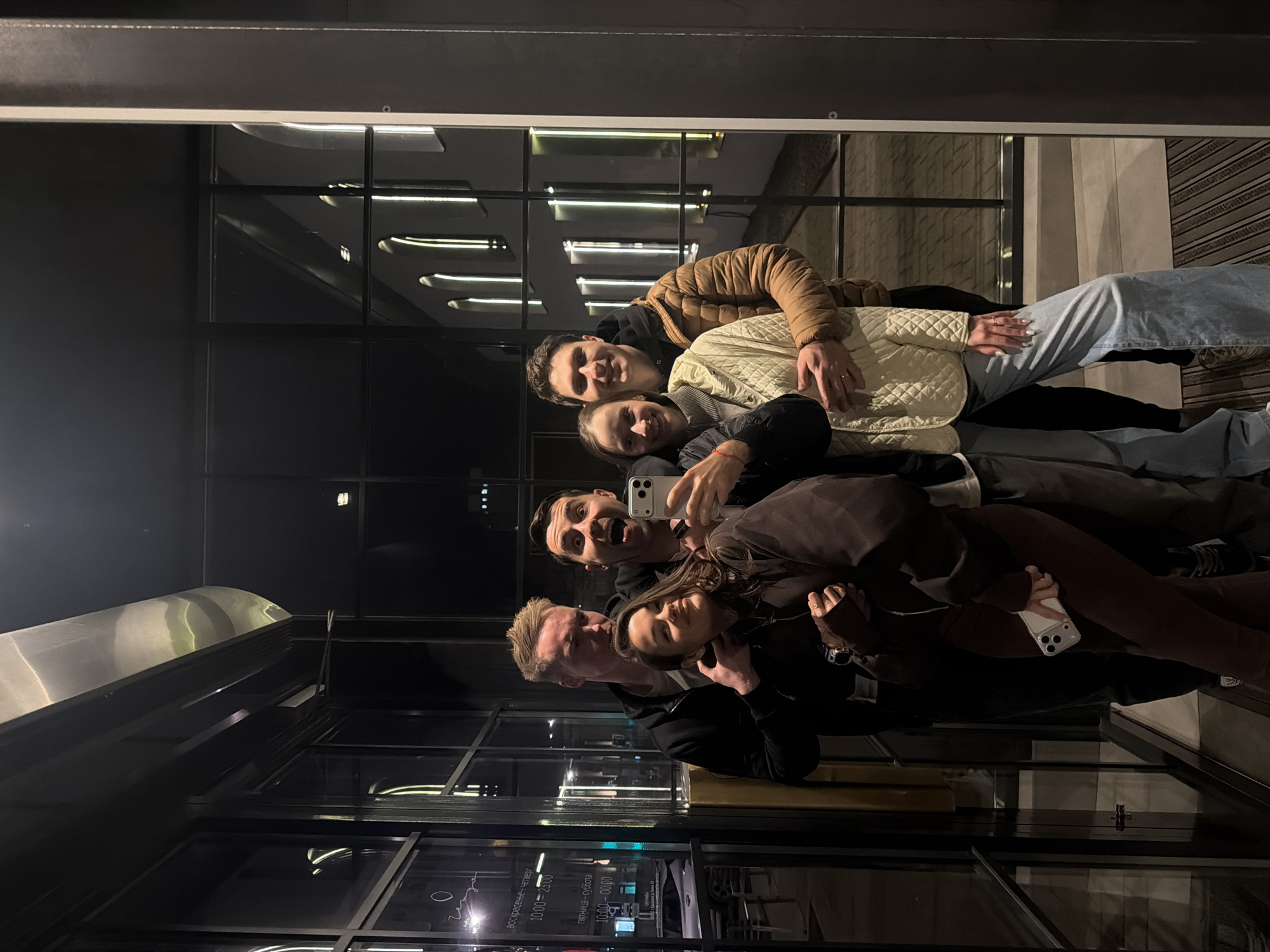 A group of six young adults poses for a mirror selfie indoors, likely in a cafe or restaurant, during the evening or night. The image is taken in Krasnoyarsk, Russia. The background features large glass panels or windows reflecting interior lights and architectural details.
The subjects appear to be in their twenties or early thirties. From left to right (as if the image were upright):
1. A man with short blonde hair, wearing a dark jacket, smiling slightly.
2. A woman with dark hair, looking at the camera and smiling, wearing a dark top and holding a white smartphone.
3. A man with dark hair, making a playful expression with an open mouth, wearing a dark top and holding a white smartphone, positioned as if taking the selfie.
4. A woman with dark hair, looking at the camera and smiling, wearing a light-colored quilted jacket.
5. A man with dark hair, smiling and wearing a brown puffer jacket over a dark hoodie, with his arm around the woman in the light jacket.
6. A woman with long dark hair, partially visible behind the man in the brown jacket, smiling faintly.
All individuals are dressed in casual, warm attire. Their expressions convey a cheerful and social mood.
In the reflection on the lower left of the image (when oriented upright), text is visible, indicating business operating hours in Russian. It reads: "ВОСКРЕСЕНЬЕ - ЧЕТВЕРГ" (Sunday - Thursday) "10.00 - 23.00" and "ПЯТНИЦА - СУББОТА" (Friday - Saturday) "10.00 - 00.00". These details reinforce the setting as a public establishment with specific operating times. The overall ambiance suggests a modern, urban environment.