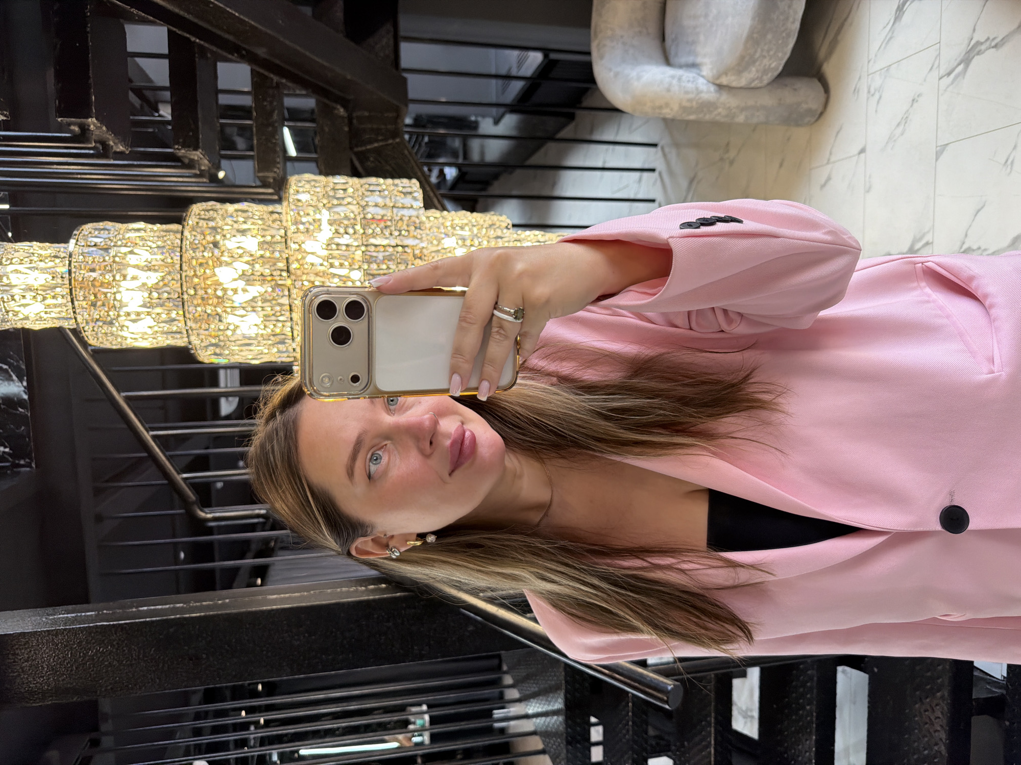 A woman is taking a selfie in what appears to be a building with a staircase, in Moscow, Russia. The woman is wearing a pink blazer. The woman is holding a smartphone in her right hand. A chandelier is in the background. The staircase has black metal railings. The building has white marble flooring. There is a sculpture in the background.