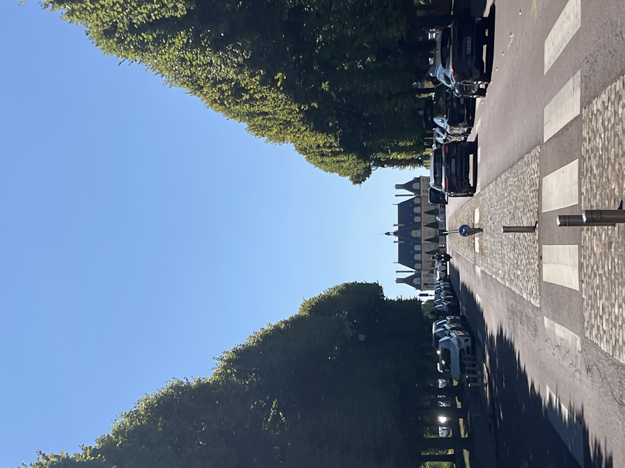 The image depicts a straight, tree-lined avenue in Sceaux, France, leading to a large, ornate building with multiple peaked roofs. The avenue is paved with cobblestones in the center and has asphalt on the sides. Cars are parked along the right side of the avenue and are driving down the center lane. Pedestrian crosswalks are visible on the cobblestone section. On the left side of the avenue, a row of manicured, tall trees creates a dense green wall. The sky is a clear blue. The building at the end of the avenue appears to be a grand estate or chateau, possibly the Palais de l'Île d'Amour, given its architectural style and the context of Sceaux.