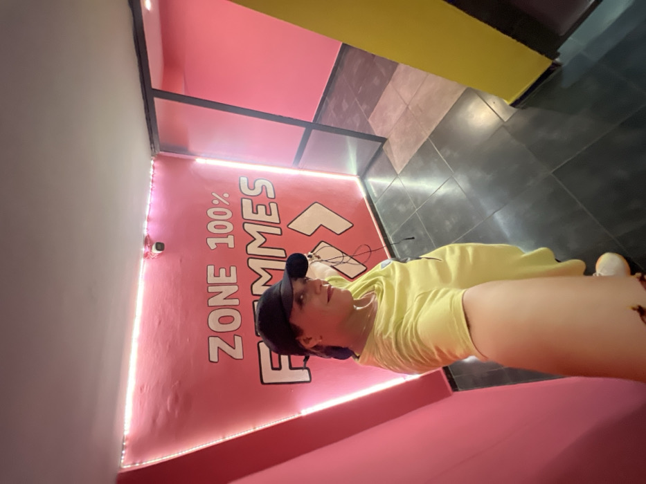The image depicts an indoor scene, possibly a brightly decorated gym or recreational space, with a person taking a selfie. The perspective suggests the person is holding the camera at arm's length, angled slightly upwards, and is positioned low in the frame, with much of the background visible above them.
The central figure is a person with light skin, wearing a bright yellow short-sleeved t-shirt and a dark-colored baseball cap. Their head is tilted slightly, and they are looking towards the camera with a neutral expression. Dark hair is visible underneath the cap. They appear to have earphones in their ears, with wires extending from them. Their right arm (from the viewer's perspective) is extended to take the photo, and part of their left arm and upper body are also visible.
The background is characterized by bold, contrasting colors and distinct architectural features. The wall immediately behind the person is a vibrant, bright pink. Large, stylized white letters are printed vertically on this pink wall, reading "ZONE 100% FEMMES" (meaning "100% Women Zone"). An arrow graphic also points upwards next to the text. Along the bottom edge of this pink wall, where it meets another surface, a prominent strip of bright white LED lights illuminates the space.
To the left of the pink wall, another wall is visible, appearing to be a light gray or off-white color. A small, white dome-shaped security camera or sensor is mounted on this light-colored wall, just above the LED strip.
Above the pink and light gray walls, there are sections of ceiling or upper walls. A black metal frame or beam structure separates these sections. Some areas are painted bright pink, matching the main wall, while others are a vivid yellow. The floor is covered with dark gray tiles, showing a grid pattern. The overall lighting is bright, especially from the LED strip, and appears to be artificial. The combination of bright colors and specific text suggests a dedicated area or facility for women.
