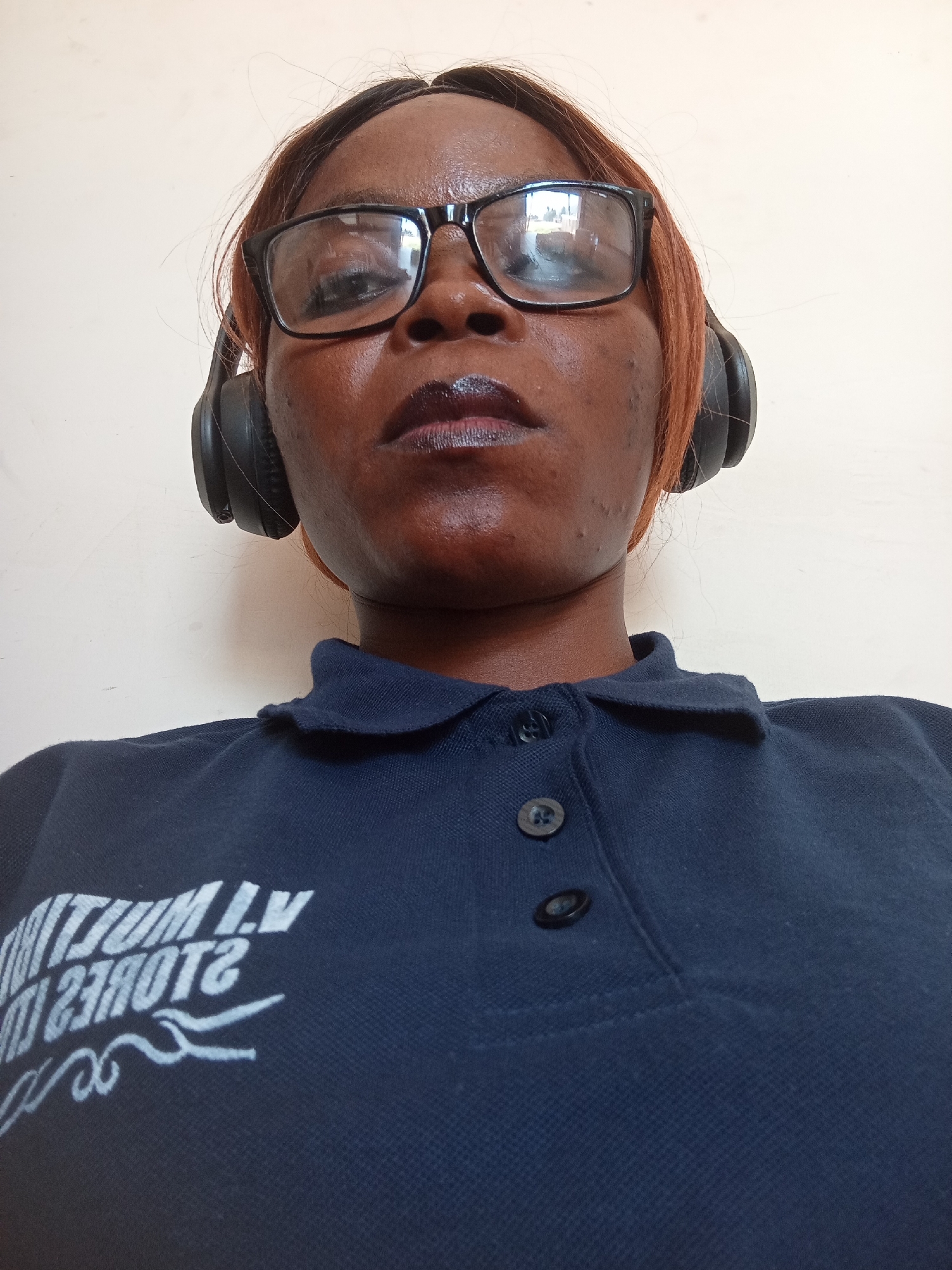 A low-angle, eye-level, medium shot captures a Black woman from the chest up, facing the camera and wearing headphones. She has dark skin, short reddish-brown hair, and is wearing black-rimmed glasses. Her lips are dark, and her complexion is lightly tanned with some visible blemishes on her chin and cheek. She is wearing a dark blue collared polo shirt with two visible buttons at the placket. White text is visible on the left side of her shirt, appearing to be a logo or inscription. The background is a plain, light-colored wall. The lighting is neutral, suggesting an indoor setting. No specific landmarks of Jalingo, Nigeria, are visible in the image.
