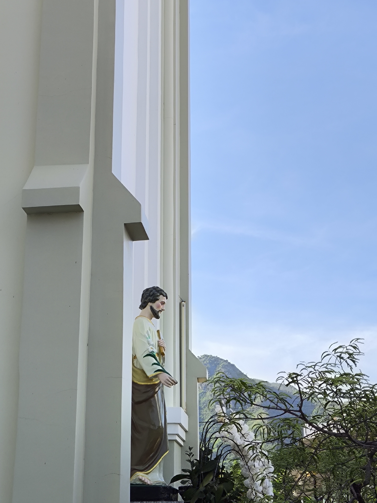 A statue of a male figure with a beard and dark hair, wearing a light-colored robe with a brown sash, stands in a niche against a white building with vertical fluting. The statue is positioned to the left of the frame, facing right, with its head tilted downwards. In the background, a clear blue sky with faint clouds transitions to green, tree-covered mountains. Lush green foliage, including a tree with delicate leaves and a branch bearing white flowers, is visible in the lower right portion of the image. The overall scene suggests an outdoor religious or decorative setting, potentially in a city like Nha Trang, Vietnam, given the architectural style and mountainous backdrop.
