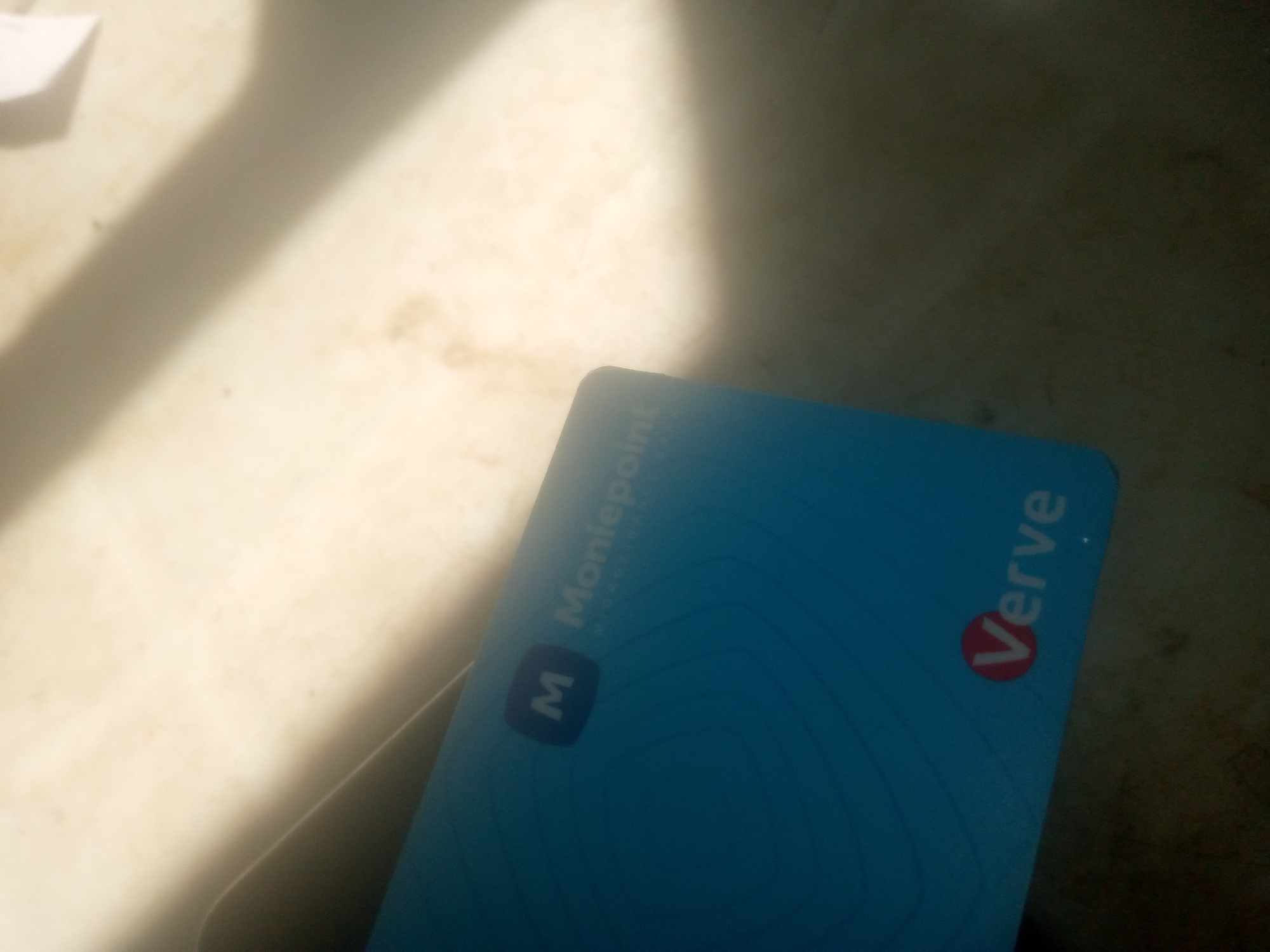 The image shows a blue Verve card with the Moniepoint logo. The card is partially obscured by shadows. The background appears to be a light-colored surface with some texture, possibly a table or countertop. The lighting suggests an indoor environment with a strong light source casting shadows.