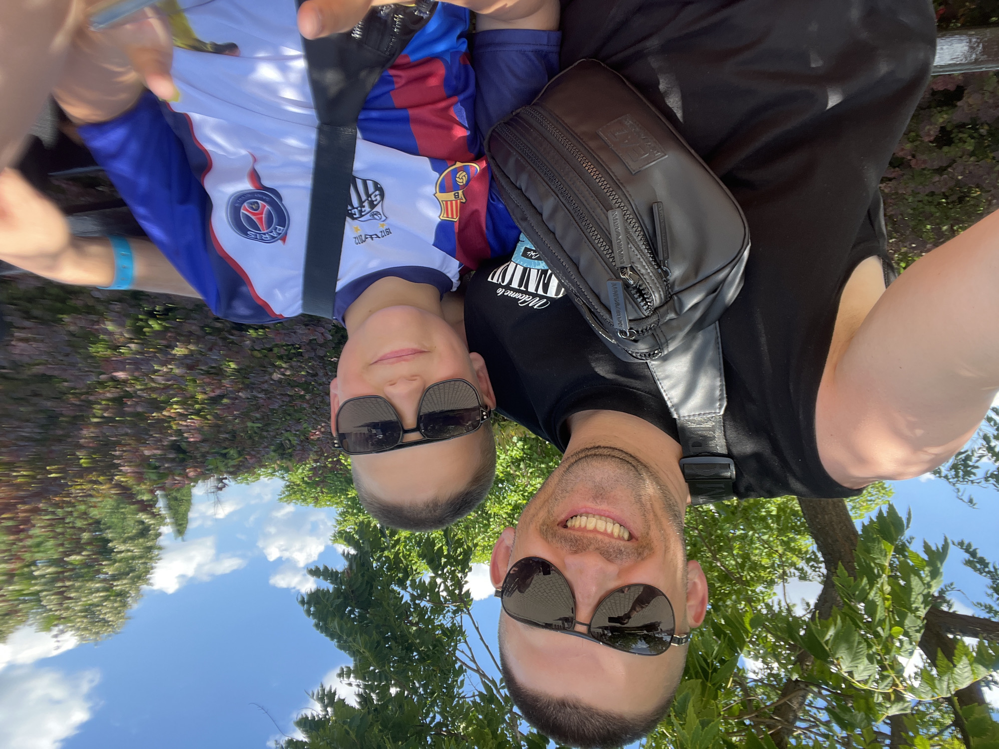 Two males, an adult and a child, are captured in a close-up, selfie-style photograph taken outdoors. The adult, on the left, wears a black sleeveless shirt and a black fanny pack across his chest. He has short dark hair, a light beard, and is wearing aviator sunglasses. He is smiling broadly, revealing his teeth. The child, on the right, wears a t-shirt with a soccer team logo, a blue collar, and red and blue striped sleeves. He also wears aviator sunglasses and is smiling. Both individuals are seated, and in the background, there are green trees, bushes, and a bright blue sky with white clouds. The setting appears to be a park or a recreational area. The image includes visual cues that place the location in Ravda, Bulgaria, specifically the park area near the beach.