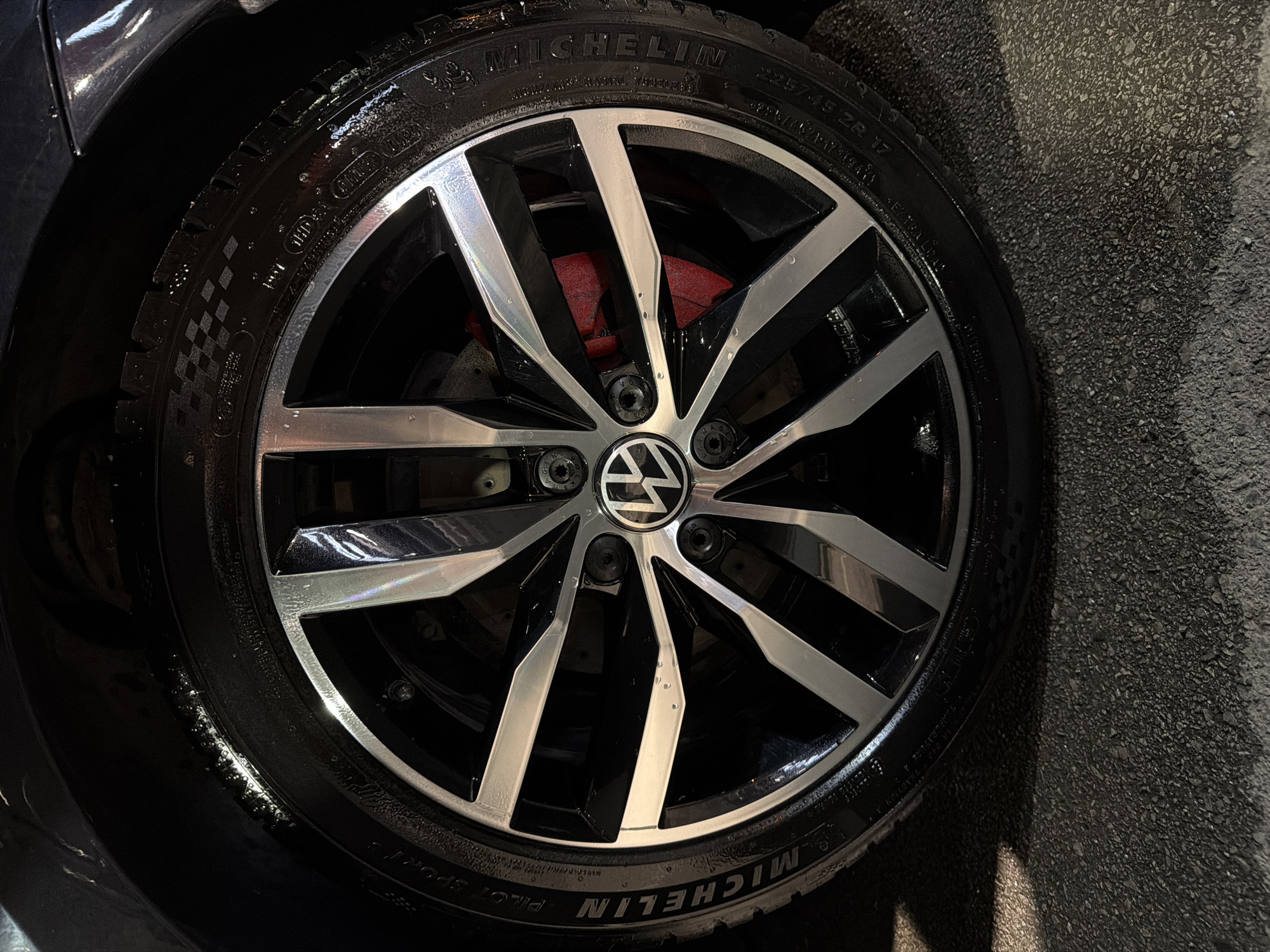 A close-up shot captures a wet car wheel, consisting of a tire and a multi-spoke alloy rim, positioned against a dark background with textured dark grey ground visible on the right. The wheel appears to have been recently washed or to have encountered wet conditions, as it is covered in numerous water droplets and glistening surfaces.
The tire is a "MICHELIN" brand, specifically from the "PILOT SPORT" series, with the size "225/45 ZR 17" clearly legible on its sidewall. Additional text like "RADIAL" and "EXTRA LOAD" can also be identified. The tire tread features a distinctive checkerboard pattern.
The alloy rim is characterized by a design of alternating polished silver and black spokes, with the silver sections having a brushed or machined finish. At the center of the rim, the circular "VW" (Volkswagen) logo is prominently displayed. Behind the spokes, a bright red brake caliper is visible, providing a contrasting color element. Small text, possibly "WAG", is faintly visible on the lug bolt covers around the central VW emblem.
The left side of the image shows a dark, reflective surface, likely part of the car's fender or bodywork. The textured ground on the right resembles asphalt or concrete, also appearing damp. The lighting suggests an indoor or covered setting, possibly in a garage or car wash in Sofia, Bulgaria. The image highlights the clean, detailed aesthetic of the car's wheel.