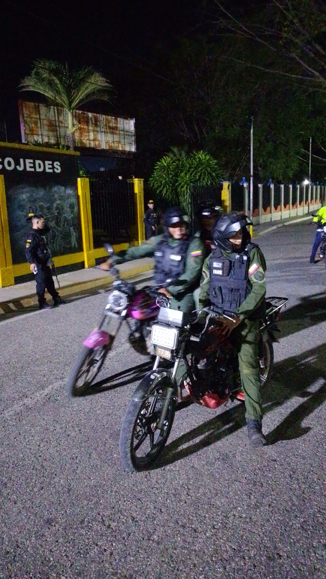The image shows a street scene at night in what appears to be the city of Tinaquillo, Venezuela, based on the sign "COJEDES" visible on a building in the background. Two uniformed personnel, possibly law enforcement or military, are seated on motorcycles. Both wear tactical vests and helmets or caps. One of them has a patch on the vest indicating the letters "FANB". A third uniformed person stands nearby, holding a rifle. Other uniformed individuals are visible further in the background. The area is lit by streetlights. The motorcycles are of a small, basic type. One is painted pink.