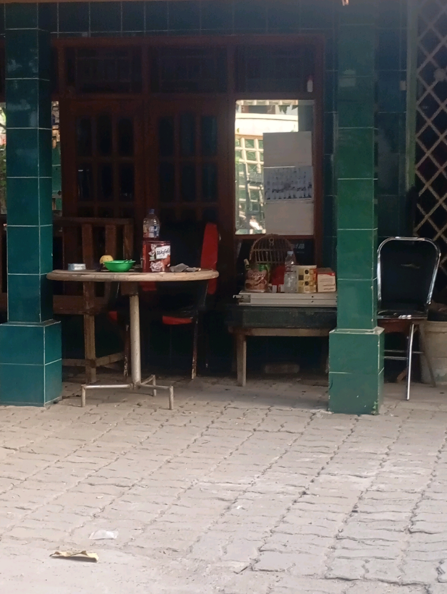 This image depicts an outdoor seating area in front of a building with wooden doors and windows in Babelan, Indonesia. A round, metal-legged table is positioned in the foreground, set with a green bowl, a yellow fruit, a can, and a bottle of water. Two chairs are visible behind the table, one with a red back and another partially obscured by a green tiled pillar. To the right of the table, a long, low table holds a birdcage, several bottles, and boxes. A black metal chair with a silver frame is situated to the far right, near a white bucket. The ground is paved with interlocking concrete blocks. The building's facade features dark green tiled pillars.