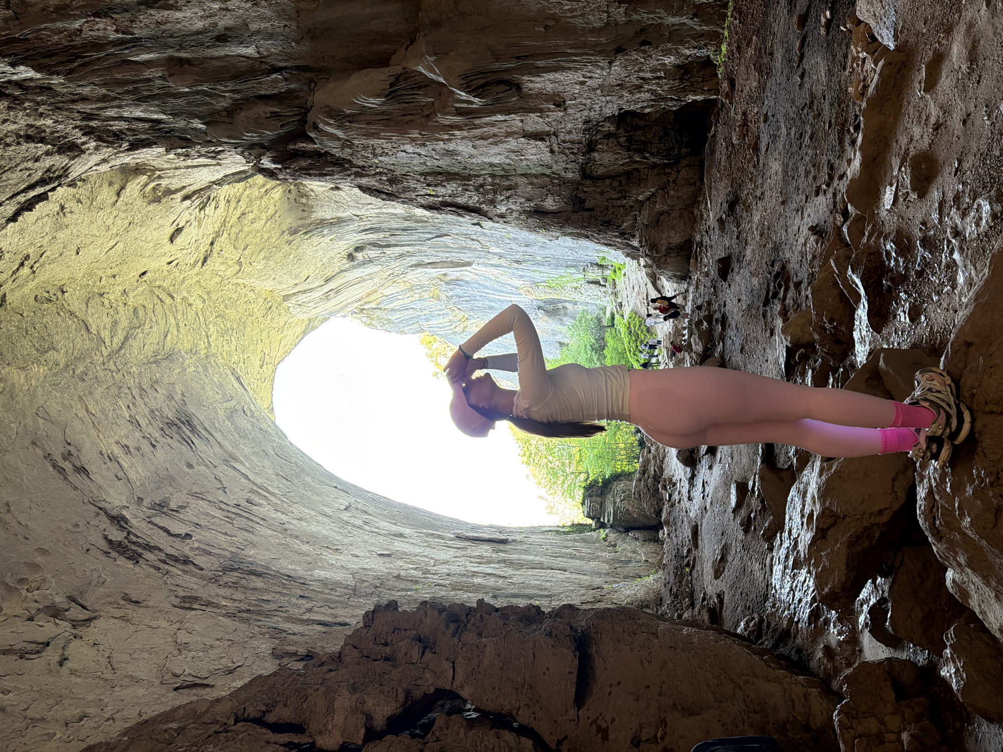 The image depicts a woman standing within Devetashka Cave, located near the city of Karlukovo, Bulgaria. She is wearing light-colored, tight-fitting clothing and pink accessories (hat, socks). She has her hands raised towards her head, possibly adjusting the hat. The cave's entrance, a large oval opening, is visible in the background, allowing natural light to illuminate the scene. The cave's interior walls are rocky and textured, with some areas appearing damp. In the background, near the exit, a group of other people are present.