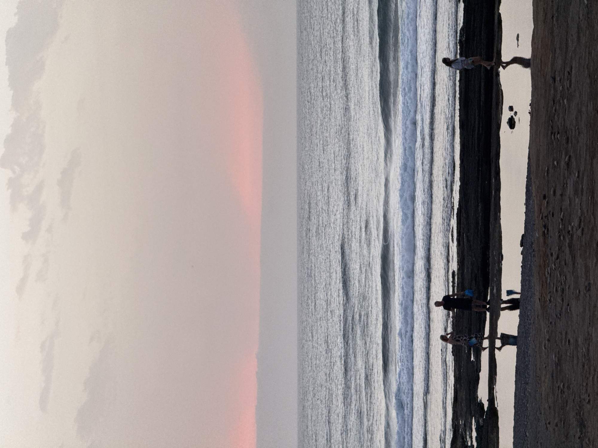 A wide-angle, full shot captures a beach scene at dusk in Arona, Spain. The sky is a soft gradient of pale pink and beige, with a thin band of darker clouds near the horizon, where a subtle pink glow indicates the setting sun. Below the sky, the ocean stretches out, calm with gentle waves breaking on the shore. The foreground is a dark, rocky shoreline with tidal pools reflecting the muted sky. Several silhouetted figures are present. Two individuals, a man and a woman, stand near each other on the rocks, both holding blue bags. Further to the right, a lone woman walks along the rocky area, her reflection visible in a tidal pool. The beach itself in the very bottom foreground is a dark, pebbly sand.