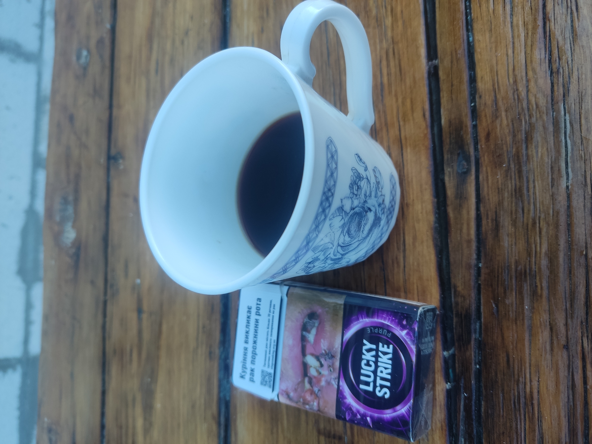 A pack of Lucky Strike Purple cigarettes and a white mug filled with dark liquid sit on a weathered wooden surface. The cigarette pack displays a graphic warning image and text in Ukrainian. The mug is decorated with blue ornamental patterns. In the background, a blurred brick wall is visible. The scene is outdoors, likely on a table.