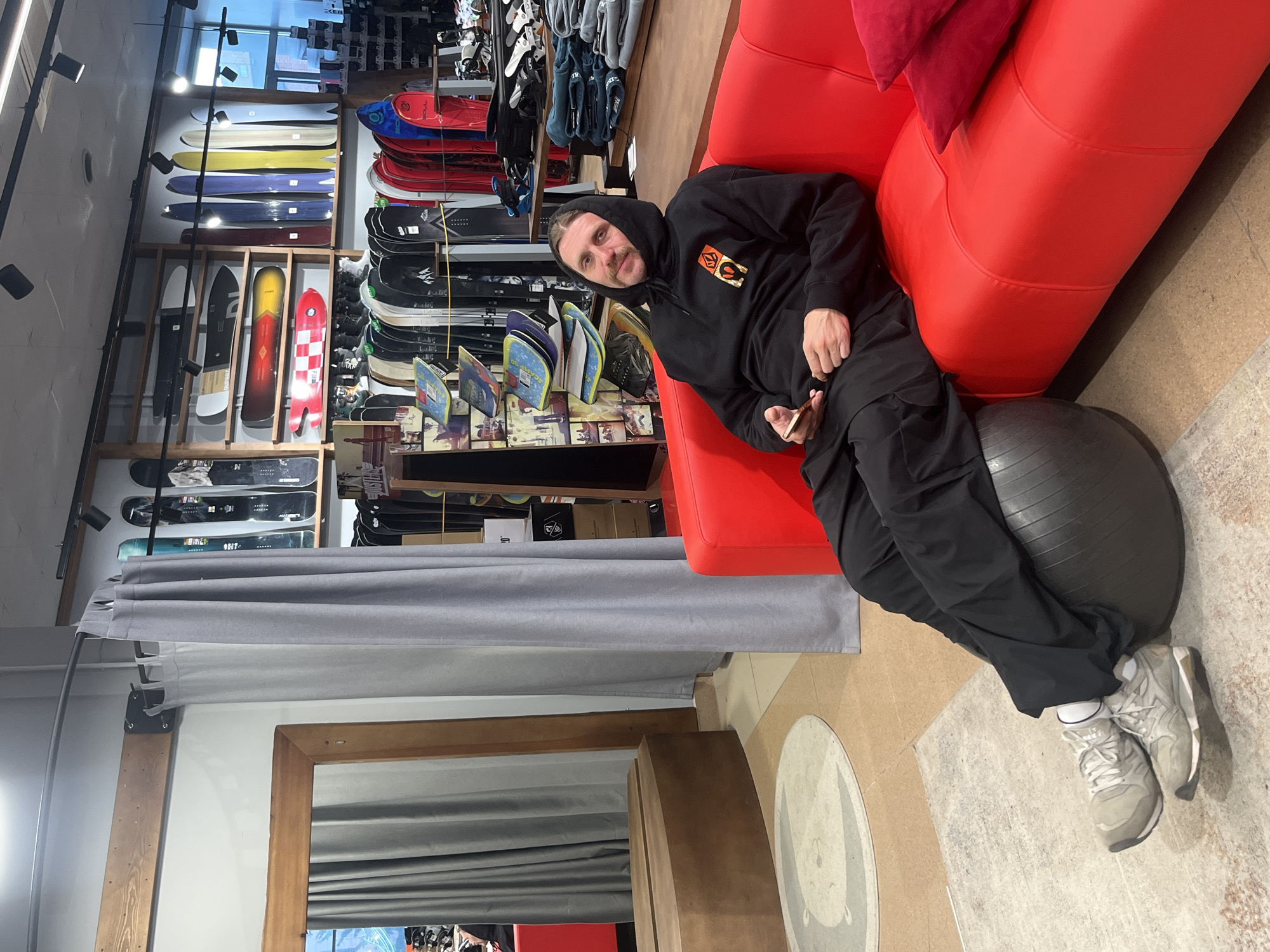A man is casually seated on a bright red sofa, leaning back, with his feet resting on a large black exercise ball. He is wearing a black hooded sweatshirt with an orange and yellow logo on the chest, black pants, and light grey sneakers with white socks. He holds a smartphone in his right hand, looking directly at the camera with a neutral expression.
The setting is an indoor retail store, specifically a snowboarding shop, as indicated by numerous snowboards displayed on wooden and metal racks behind the man. The snowboards vary widely in color and design, featuring brands like Burton and Lib Tech visible on some. Other merchandise, including bindings and possibly apparel, is also visible. The store has a mix of flooring, with light brown and grey textured sections, and a circular rug with a bear outline is partially visible near the man's feet. Light grey curtains hang along one wall, and a wooden-framed mirror is seen at the bottom. The lighting suggests an indoor commercial environment. The overall atmosphere appears relaxed, with the man seemingly taking a break or posing in the shop located in Krasnoyarsk, Russia.