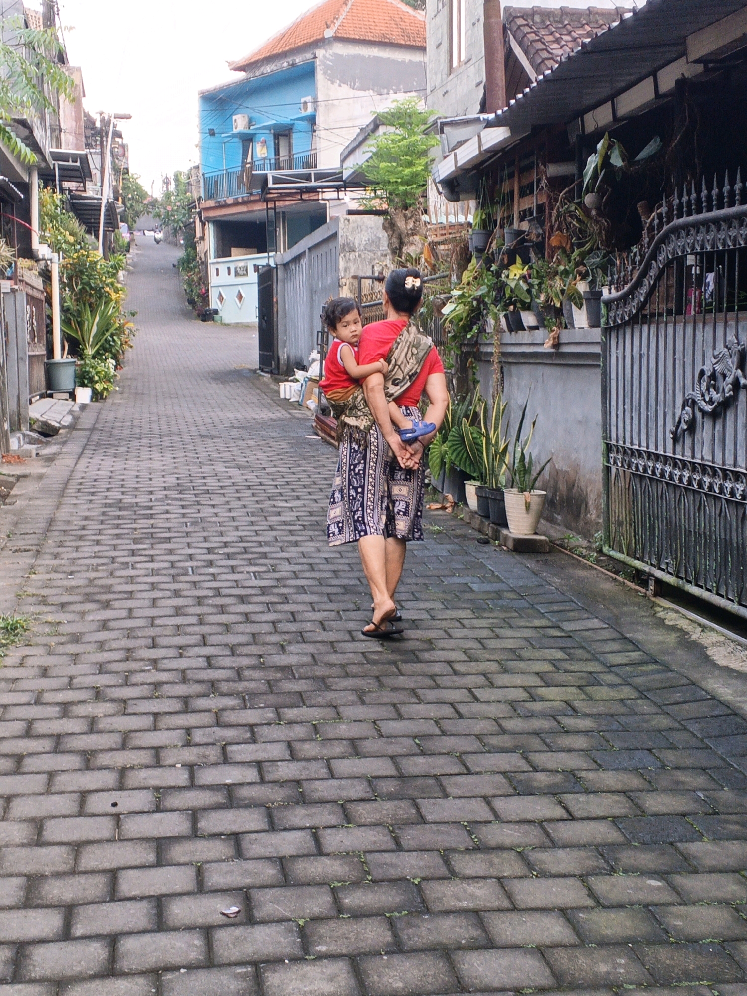In an alleyway in Denpasar, Indonesia, a woman is seen from behind, carrying a young child on her back, who is wrapped in a sarong. The woman wears a red top and a patterned skirt, with sandals on her feet. The child, also in a red top, looks over the woman's shoulder. The alleyway is paved with grey bricks and lined with buildings, some of which are colorful. Lush greenery, including potted plants and climbing vines, adds a vibrant touch to the scene. A decorative metal gate and fence are visible on the right side of the image. The overall atmosphere suggests a typical day in a residential area.