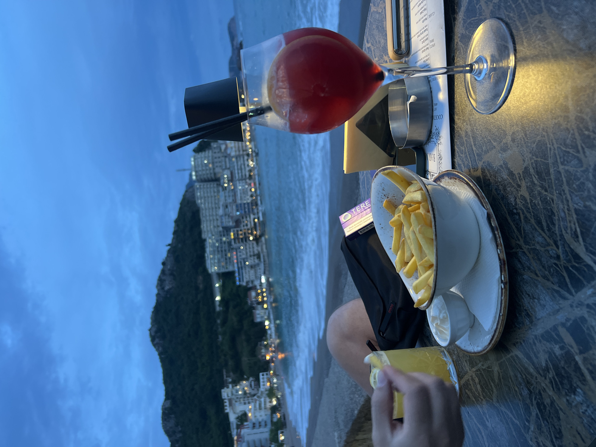 The image depicts a table setting in Rafailovići, Montenegro, with a view of the Adriatic coastline. A bowl of french fries rests on a patterned plate next to a small container of what appears to be mayonnaise or a similar dipping sauce. Next to the fries, there are a wallet and a hand holding a glass of orange juice. In the background, there is a cocktail in a tall glass with a black paper cup around it, next to a menu and silverware. The ocean stretches out into the distance, with buildings and a mountain visible along the shore. The sky is a dusky blue, suggesting either dusk or dawn.
