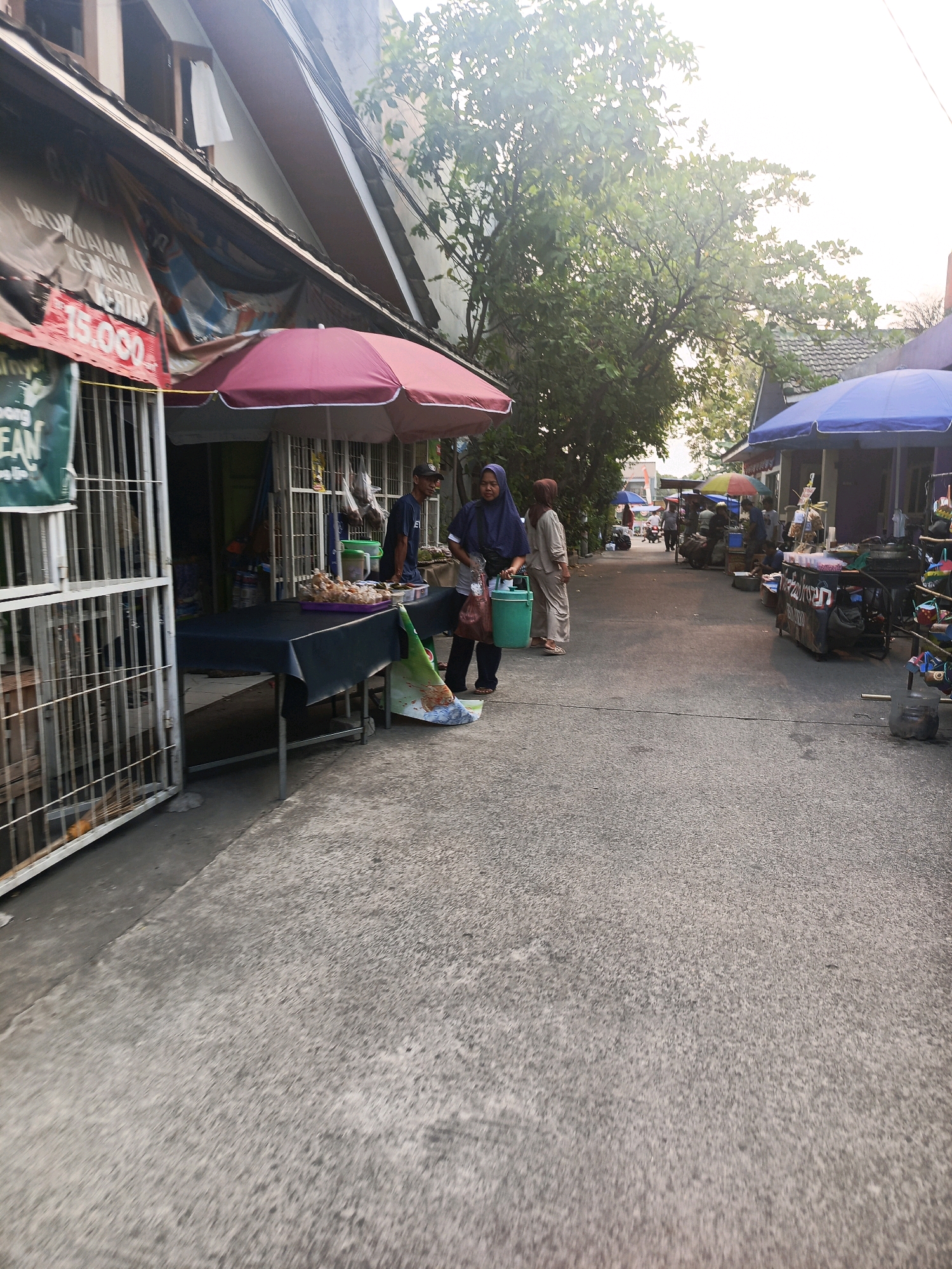 This image depicts a street scene in Balaraja, Indonesia, likely a marketplace or a commercial street with various stalls. In the foreground, a concrete pathway is visible, with a building on the left side featuring a prominent red sign displaying "15.000". Adjacent to this building is a market stall covered by a burgundy umbrella, with a black tablecloth laid out on a table laden with food items, likely snacks or pastries. A person wearing a blue headscarf and dark clothing is standing at the stall, holding a green bucket and a shopping bag. Another person, wearing a light-colored headscarf and matching outfit, stands nearby.
Further down the street, on the right side, several other market stalls are visible, some with blue and yellow umbrellas, and one with a sign featuring red and black text. These stalls appear to be selling various goods, and some individuals are seen browsing or working at these stalls. A few motorcycles are parked along the street. The environment is semi-urban, with buildings lining the street and trees providing some greenery. The lighting suggests it might be late afternoon or early morning, with a bright sky and some shadows cast on the ground. The overall impression is a busy, informal commercial area.