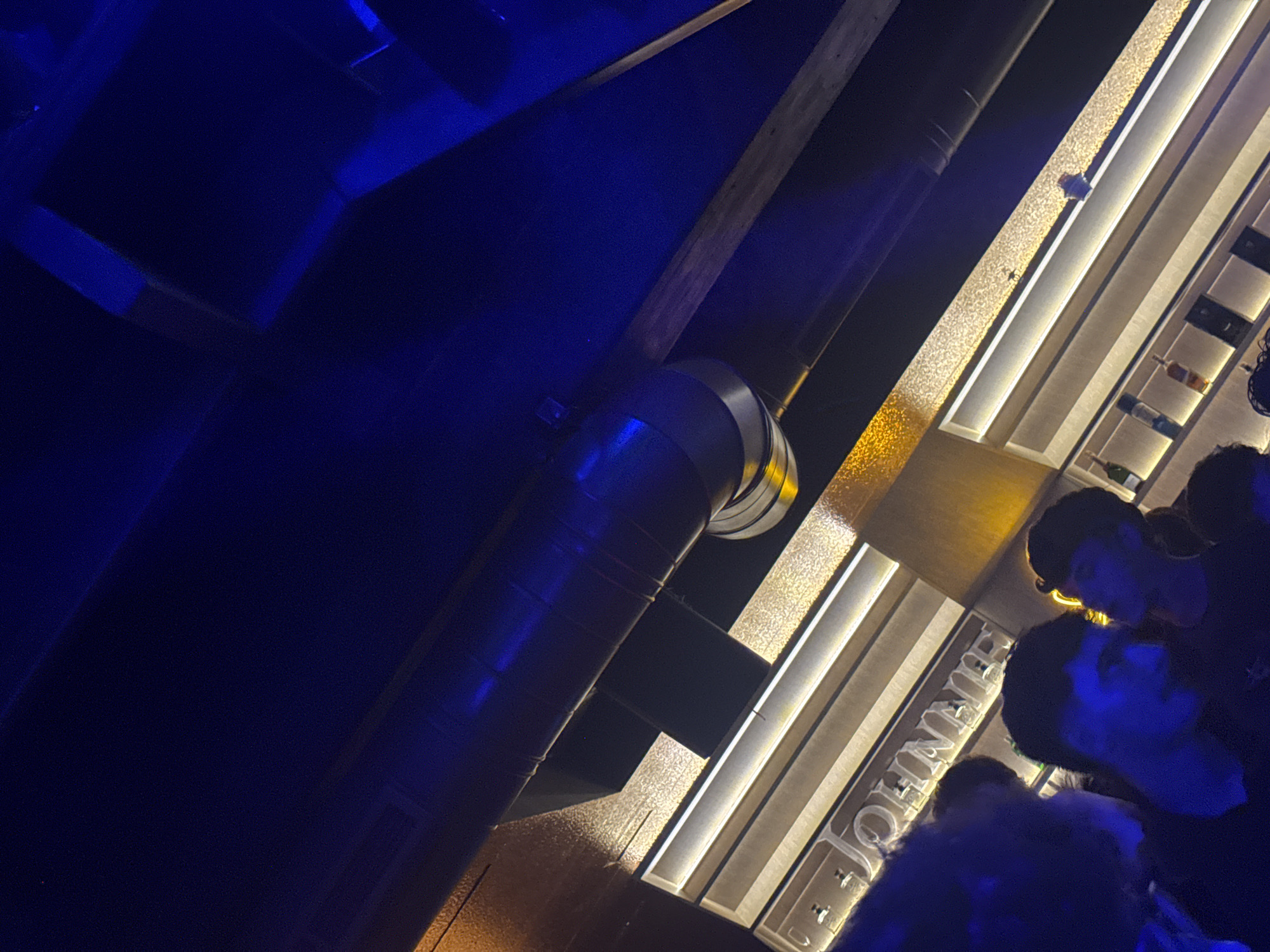 Here's a detailed description of the image:
**Environment:** The image appears to be taken inside a bar or nightclub with a modern, industrial-chic aesthetic. The lighting is dim with strong blue and yellow/gold accents. The scene features a bar with a mirrored back showcasing liquor bottles. There's exposed ductwork on the ceiling.
**Objects:** Key objects include:
* Bar counter with the word "Johnnie" visible
* Liquor bottles displayed on shelves
* Mirrored back bar
* Exposed ductwork
* Decorative lighting
**People:** There are several people present near the bar. It's difficult to discern their actions clearly, but they appear to be patrons. Their faces are partially visible, with strong blue lighting affecting their appearance.
**Actions/Interactions:** It's difficult to determine any specific actions or interactions due to the limited visibility and angle.
**Location (Inferred):** While the image does not explicitly provide information regarding Caracas, Venezuela, there are indicators that this is likely the location. The "Johnnie" signage on the bar aligns with the popular "Johnnie Walker" brand which is available in many parts of Venezuela.
**Forensic Summary:** The image depicts a low-lit interior scene, likely a bar or nightclub in Caracas, Venezuela, identified by the popular "Johnnie" brand on the bar counter. It presents patrons near the bar.