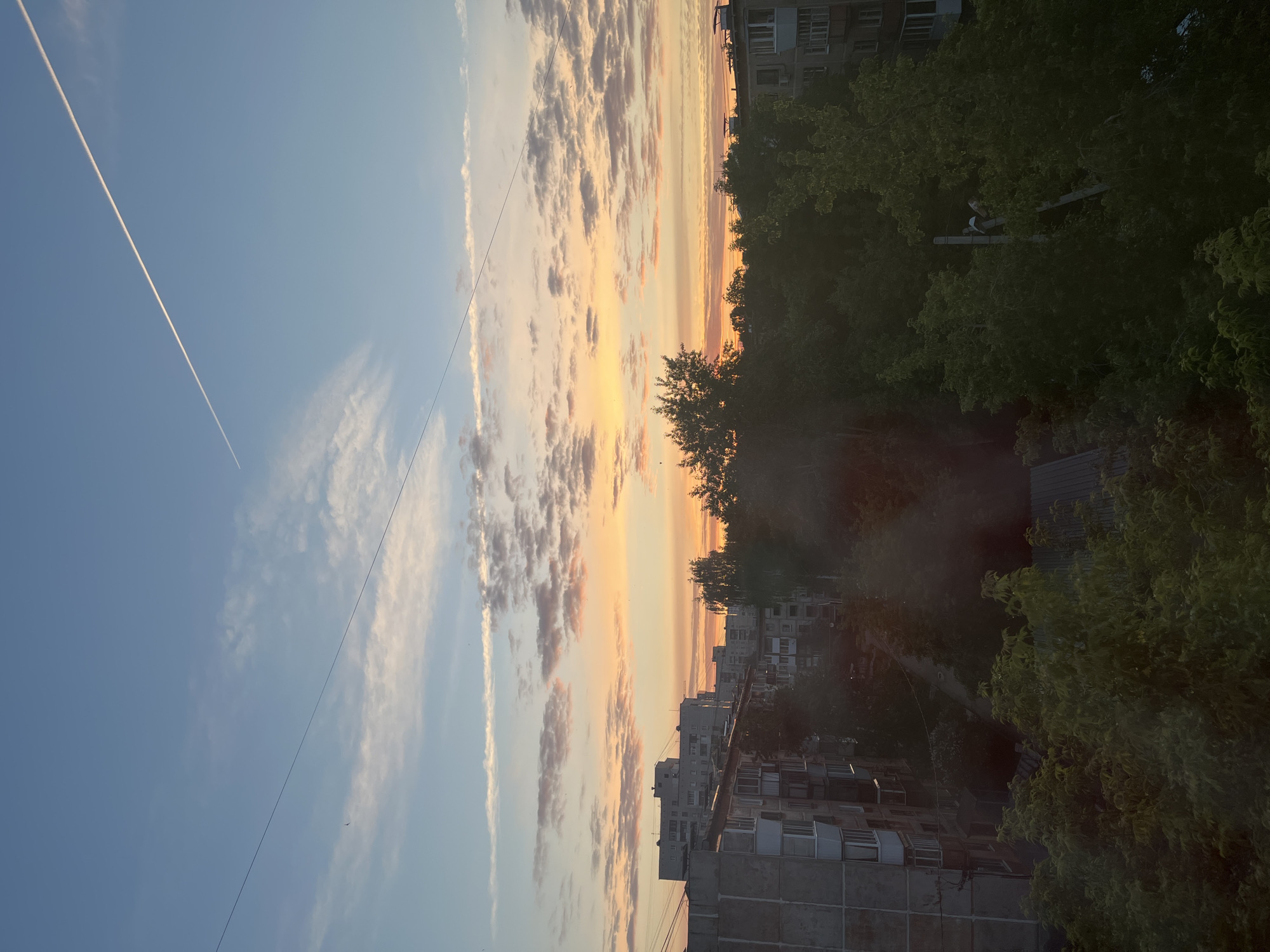 This image captures an urban or suburban landscape during either sunrise or sunset, viewed from an elevated perspective, possibly a window or balcony. The scene is dominated by a vibrant sky and a silhouetted foreground of buildings and trees.
The sky in the upper left portion is a clear, pale blue, traversed diagonally by a long, straight white airplane contrail. Moving towards the center and right, the sky explodes with warm, rich colors of orange, yellow, and pink, reflecting the low sun. These hues illuminate scattered clouds, creating a dramatic and serene backdrop. A thin, dark utility wire, likely a power or communication line, also stretches across the sky.
Below the horizon, the scene is largely in shadow. Dense, green trees with abundant foliage create a lush canopy that partially obscures the residential buildings behind them. The buildings appear to be multi-story apartment blocks, characterized by numerous windows and a somewhat uniform, functional architectural style, suggesting an older, possibly post-Soviet, urban environment. Partially hidden within the dense tree cover, some utility equipment, possibly transformers or junction boxes, are vaguely discernible.
The overall atmosphere is peaceful and calm, highlighted by the tranquil beauty of the sky during the "golden hour." There is no visible human activity or discernible text in the image.