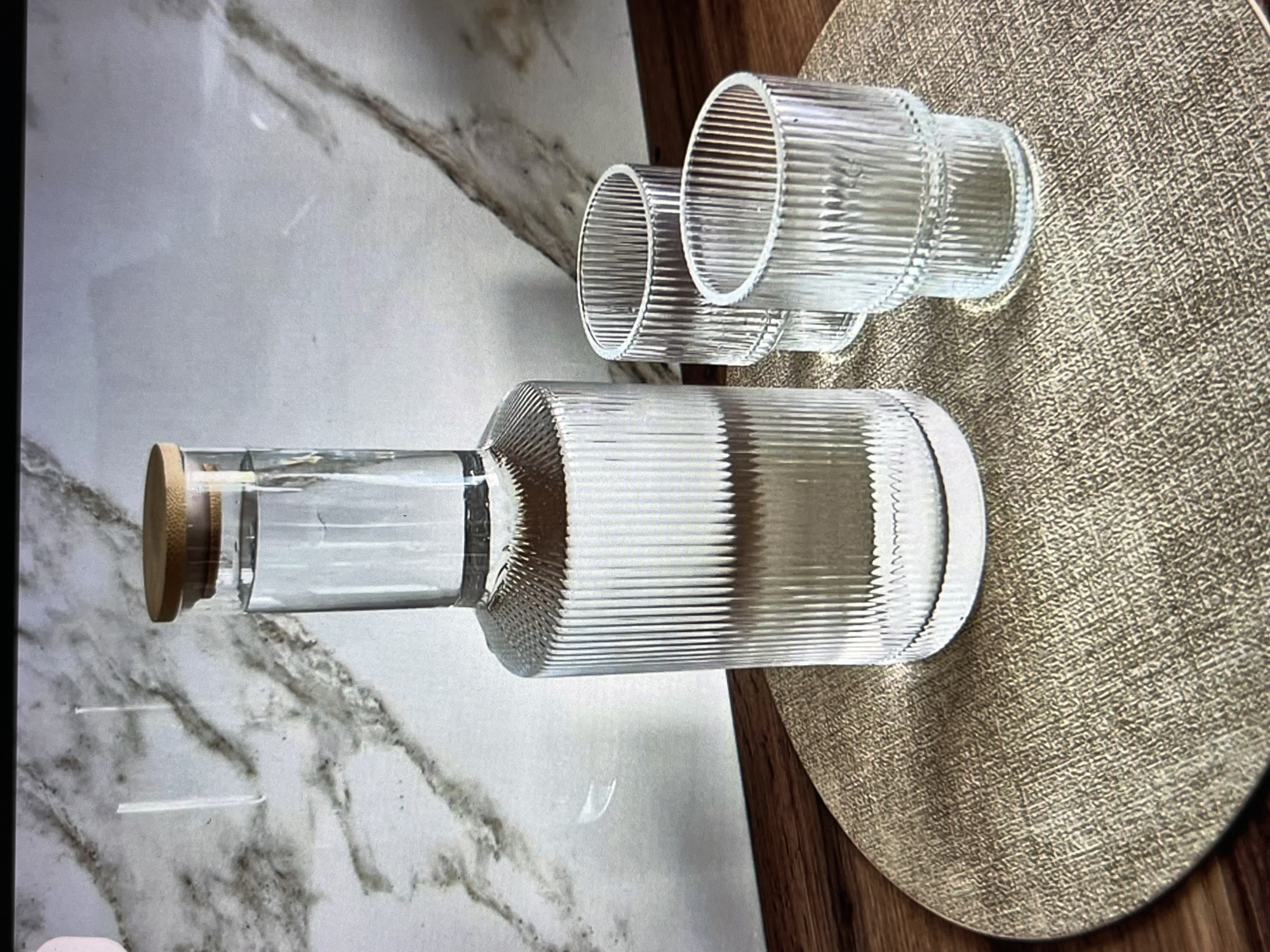 A clear glass water carafe with a wooden lid and two matching tumblers are arranged on a textured, round, gold-colored placemat. The placemat rests on a dark wood surface. Behind the placemat and subjects is a light-colored marble-patterned backsplash. The lighting suggests an indoor setting. No people are present in the image, and no specific location in Dubai, United Arab Emirates can be identified from the visual information provided.