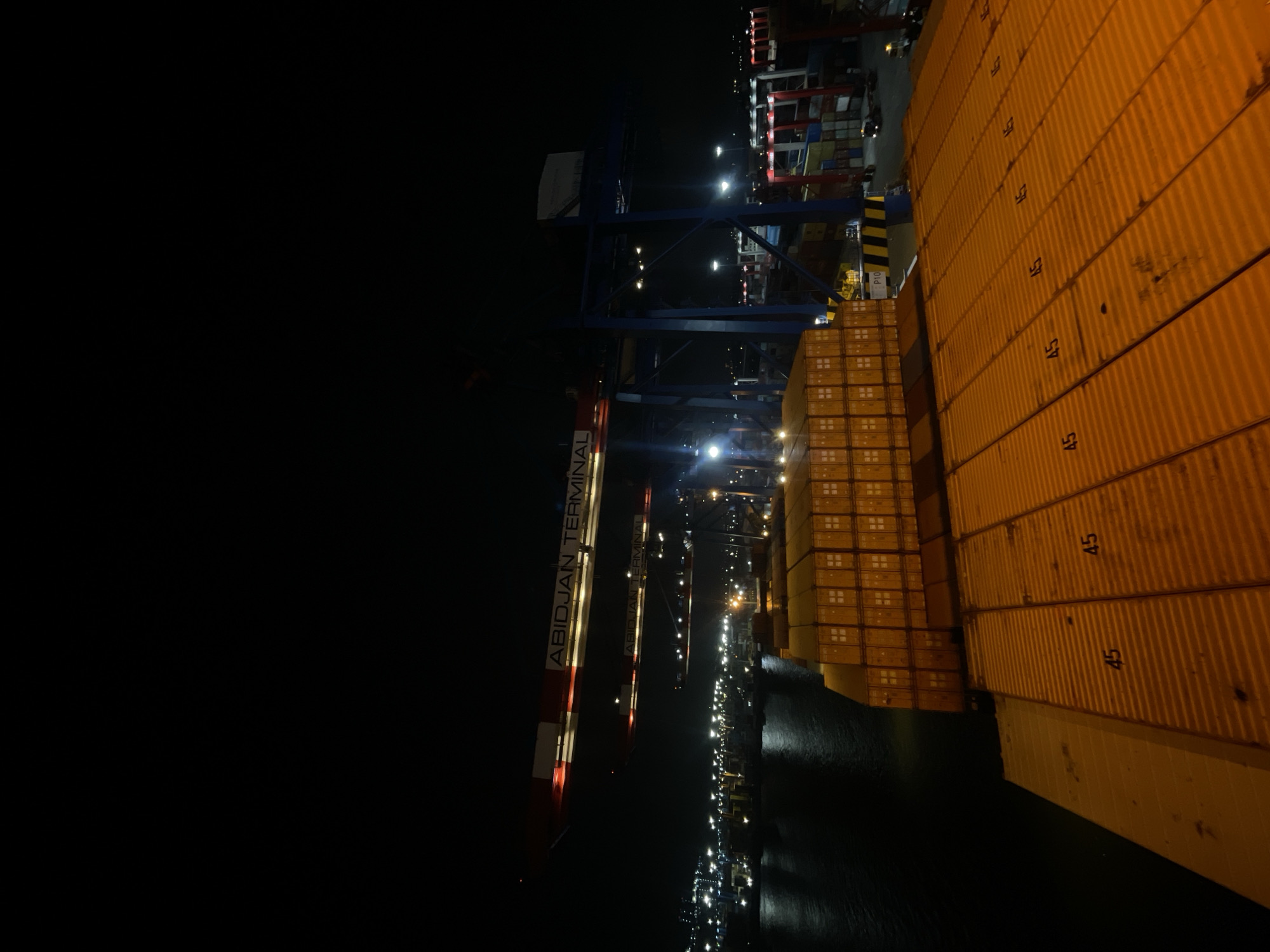 The image depicts a nighttime scene at the Abidjan Terminal, a port facility located in Abidjan, Ivory Coast. The foreground is dominated by the deck of a ship, covered in stacked orange shipping containers. Some of these containers have the number "45" stenciled on them. To the left, dark water is visible, reflecting the lights of the port.

In the mid-ground, large gantry cranes, common in port operations, are visible. These cranes are illuminated by the port's lighting system. One crane has signage that reads "ABIDJAN TERMINAL". Further back, rows of shipping containers are stacked, also illuminated by artificial lights, creating a grid-like pattern across the port. The background shows the general urban and industrial landscape of the port at night, with numerous lights from buildings and other port infrastructure. No people are clearly visible in the image. The overall atmosphere is industrial and operational, characteristic of a busy port during nighttime hours.