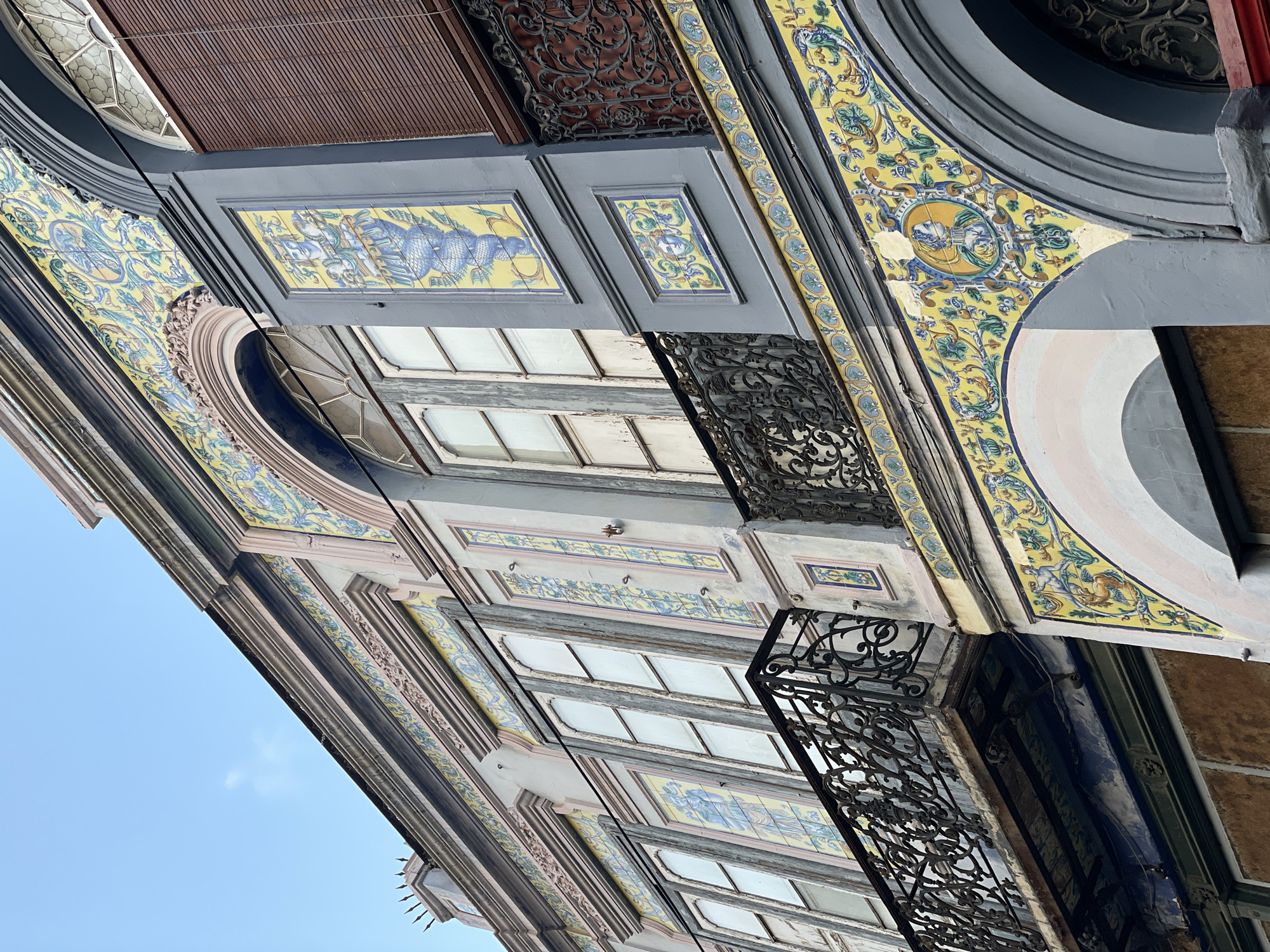 The image depicts an upward-angled view of ornate building facades in Santa Cruz de Tenerife, Spain. The architecture features a mix of grey and pastel-colored facades adorned with yellow and blue tile work depicting animals and floral designs. The building has windows and decorative black wrought iron balconies. The sky is visible in the lower-left corner, indicating the angle of the shot.