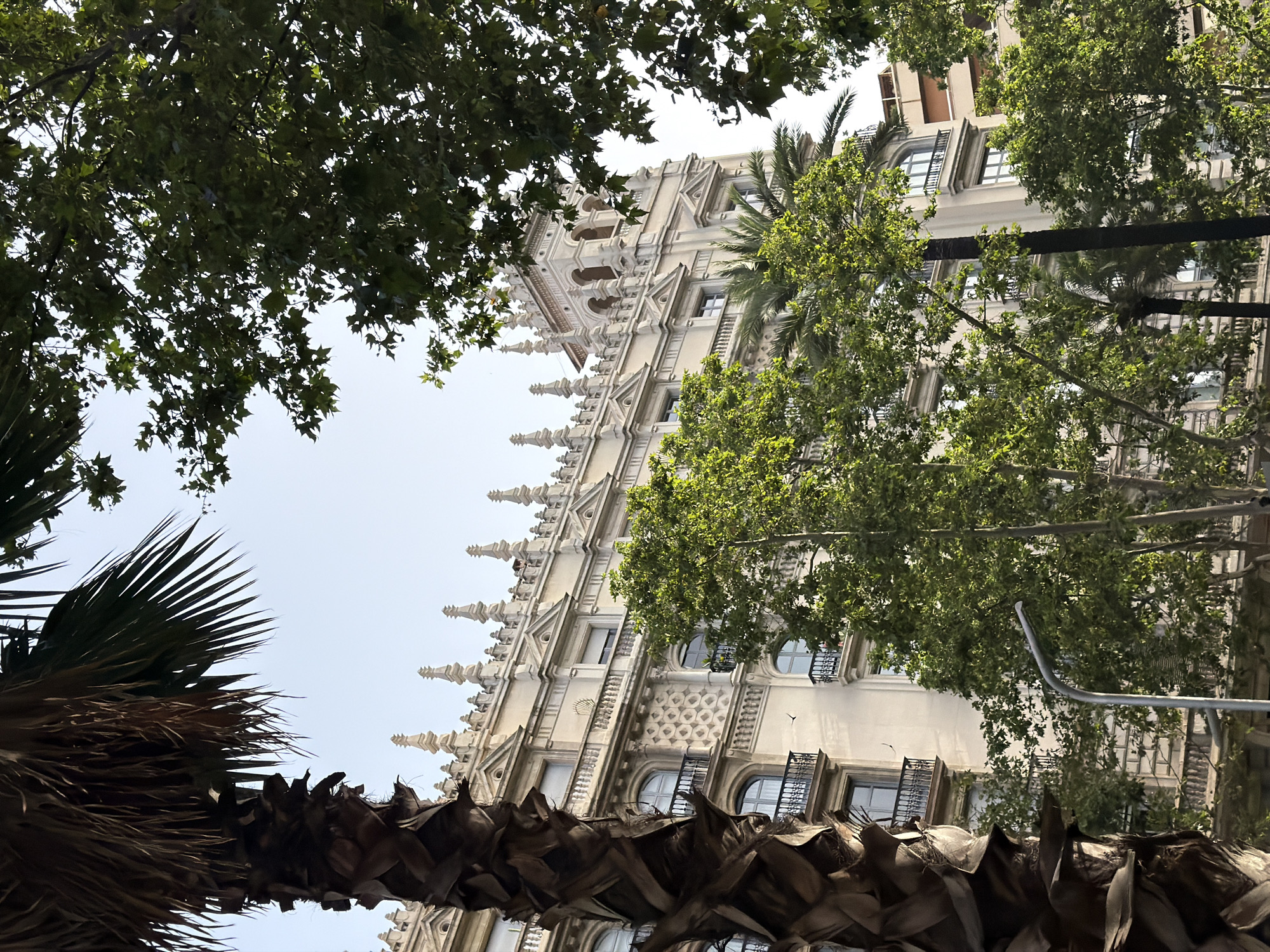 In Barcelona, Spain, a grand, ornate building with multiple floors and balconies is framed by lush green trees. A tall palm tree with a rough, textured trunk stands prominently in the foreground on the left, its fronds reaching towards the pale sky. The building features intricate architectural details, including decorative cornices, arched windows, and small spires along its rooftop. Several windows have black wrought-iron railings on their balconies. The trees, with their dense foliage, create a dappled effect of light and shadow on the building. In the lower-middle section of the image, a modern, curved streetlamp is visible, suggesting an urban setting. The sky is clear and bright, indicating a daytime scene.