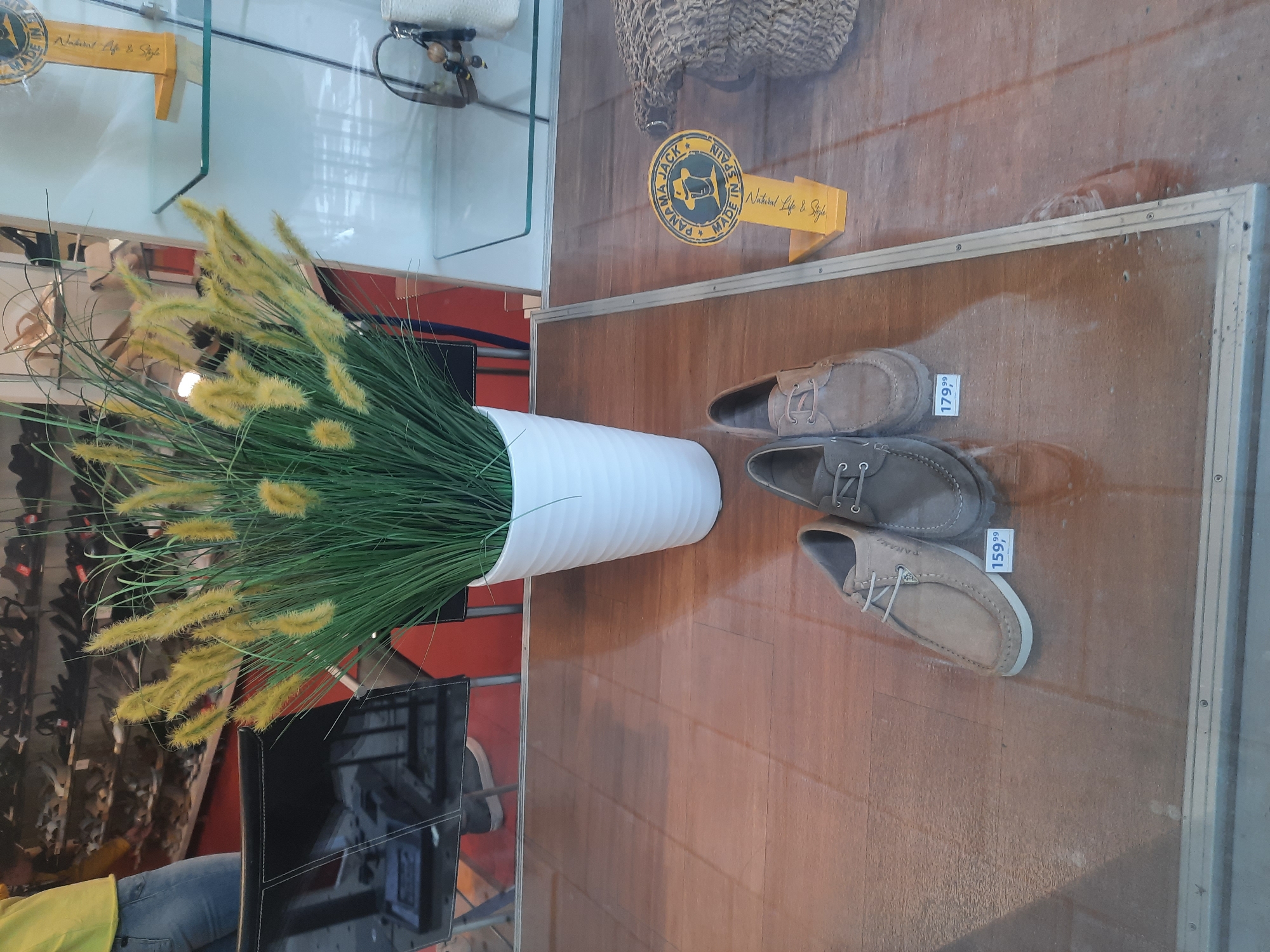 The image shows a display in what appears to be a shoe store. In the foreground, three pairs of men's shoes are arranged on a wooden surface. Two pairs of shoes are closer to the viewer. The leftmost pair is a light brown suede loafer with white laces, priced at 159.99. To its right is a darker grey suede loafer with grey laces, priced at 179.99. Partially visible behind these two is a third pair of shoes, also a light brown suede loafer.
Behind the shoes, a tall, white, ribbed planter holds a large bunch of artificial yellow grass. The planter is placed on the wooden surface.
In the background, through a glass display window, shelves stocked with shoes are visible. The lighting suggests an indoor environment. To the right of the artificial plant, a yellow sign with "PANAMA JACK MADE IN SPAIN" and a circular logo is displayed. Further to the right, a woven textured item is visible.
The scene is set within a store, with reflections visible in the glass of the display. There is no specific information in the image that places it within the city of Rosenheim, Germany; this detail appears to be external information.