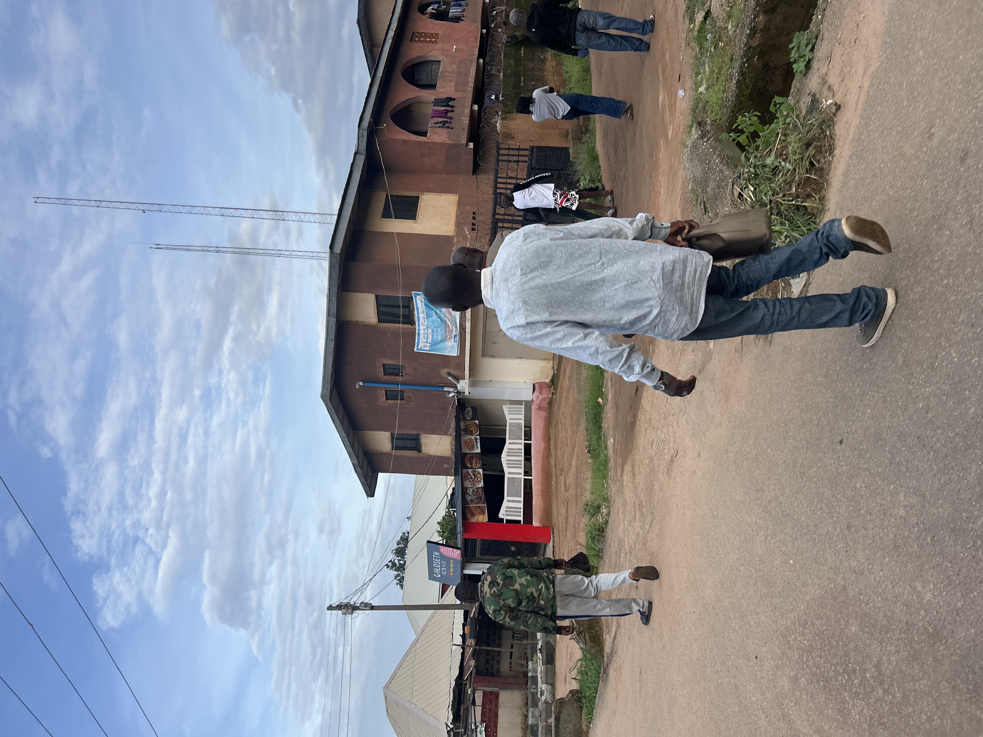 The image depicts an outdoor scene in Jos, Nigeria. The primary subjects are several people walking on a paved road. One individual in the foreground, seen from behind, is wearing a light grey long-sleeved shirt and dark blue jeans. They are carrying a brown bag and appear to be walking away from the camera. Further down the road, another person in a camouflage jacket and grey sweatpants is also walking away. Several other individuals are visible in the background, some near a building.
The setting includes a two-story building with brown and cream-colored walls, featuring arched windows and balconies with clothes hanging on a line. Adjacent to this building are smaller structures that appear to be shops, with displays visible. A sign with "CLOSSET ICE & ICE" is prominently displayed. In the distance, two tall communication towers are visible against a partly cloudy sky. The ground is a mix of paved road and dirt with some vegetation growing along the edge.
The activity appears to be casual pedestrian movement along a street. The time of day seems to be late afternoon or early evening, given the quality of light. The weather appears to be temperate with scattered clouds. The overall mood is neutral, capturing everyday life. The presence of communication towers and the architectural style of the buildings suggest an urban or semi-urban environment in Nigeria.