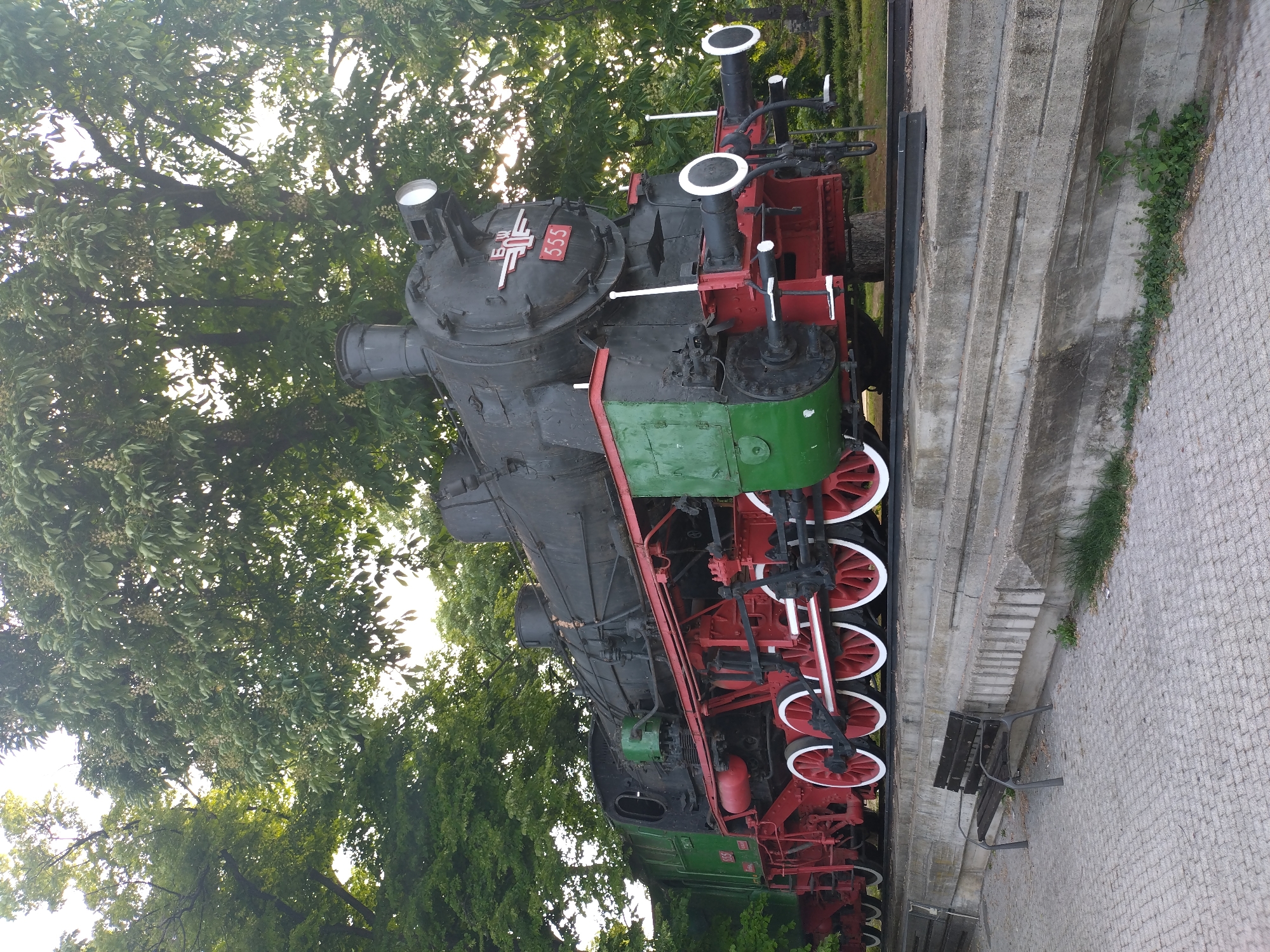 The image shows a large, vintage steam locomotive on display. The train engine is primarily black with red and green accents. The number "555" is prominently displayed on the front of the train above a white and red symbol. The wheels are red with white trim. The train is positioned on a short segment of railroad track, which is elevated slightly from the surrounding ground by a series of concrete steps. To the side of the train is a simple wooden bench placed near the steps. The scene is outdoors and surrounded by trees with dense green foliage. The locomotive's location is in the city of Stara Zagora, Bulgaria. The image captures the steam locomotive as a static exhibit within this green urban setting.