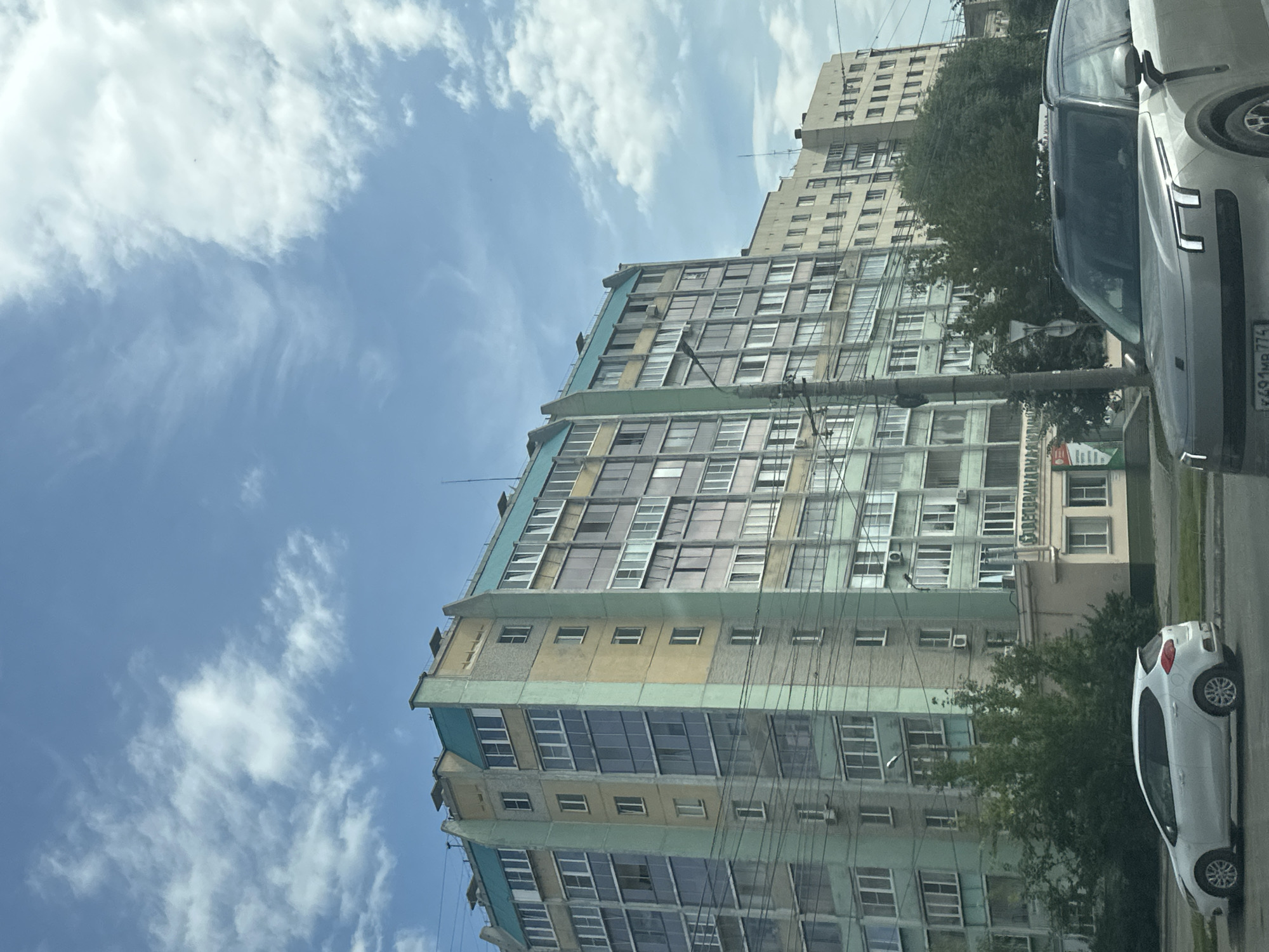 A tall, multi-story apartment building with a teal roof and beige and light green facade is viewed from street level. The building has numerous windows and balconies. In the foreground, a silver SUV is parked on the right side of the frame, and a white hatchback is parked on the left. Trees and a small building with a sign that appears to read "АПТЕКА" (pharmacy) are visible in front of the apartment building. The sky above is blue with scattered white clouds.