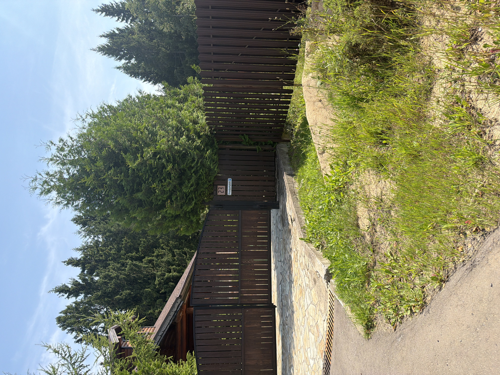 The image shows an outdoor scene in the city of Novorizhskie Klyuchi Village, Russia. A dark brown wooden fence with vertical slats encloses a property. The fence has an arched top and appears to be a gate, with two sections visible. To the left of the fence, a portion of a wooden house with a dark roof is visible. The ground in front of the fence is paved with light-colored flagstones. Beyond the flagstones and to the right of the fence, there is a grassy area with some patches of dry soil and sparse vegetation. In the background, a dense forest of tall evergreen trees can be seen. The sky is blue with a few wispy clouds. A small rectangular sign with the number "23" and some text in a foreign language is attached to the right section of the fence.
