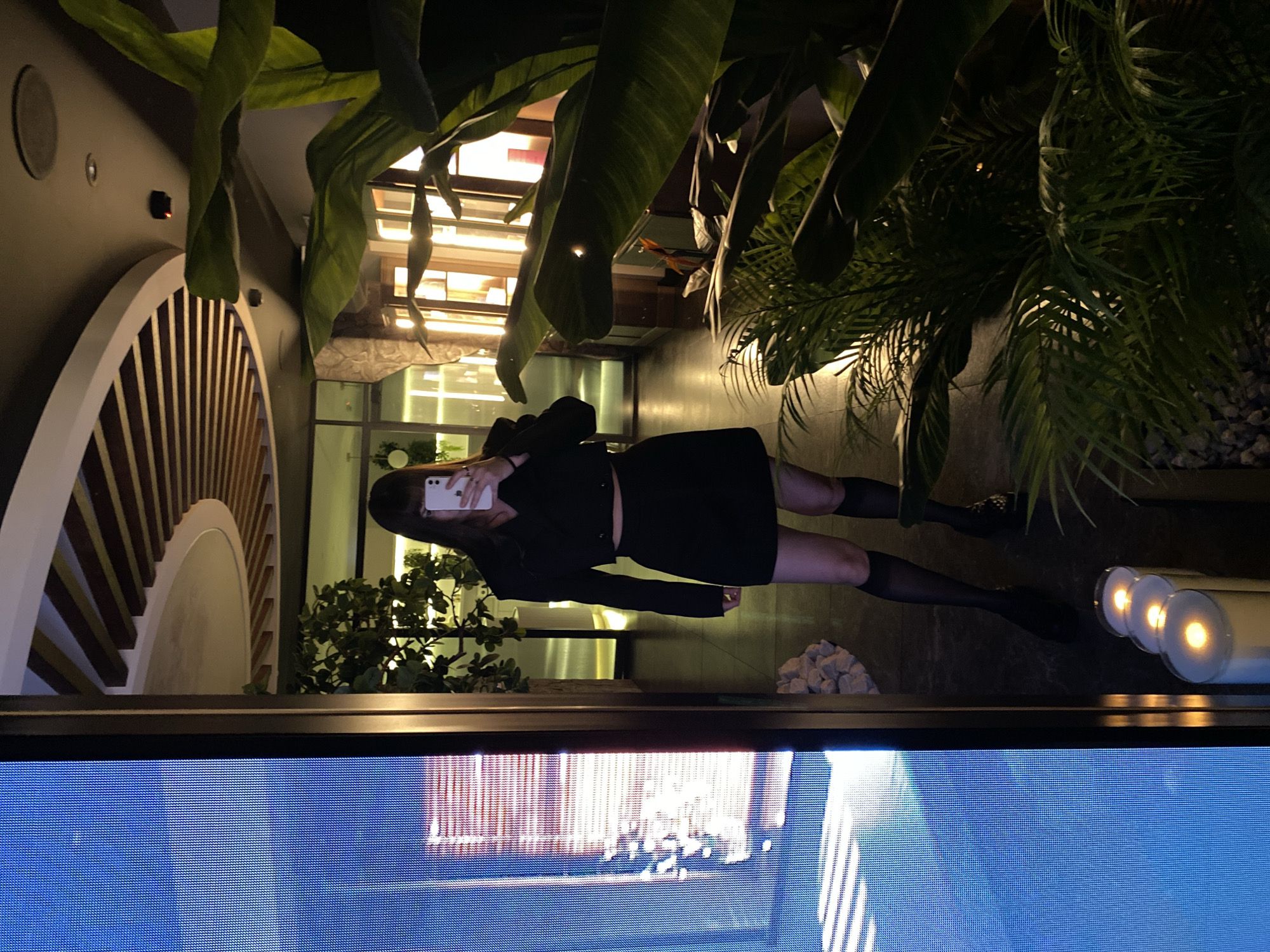 A person stands in front of a window, likely taking a picture of themselves with a phone. The setting appears to be indoors, possibly a restaurant or lounge. The area includes plants, architectural details, and illuminated features. The image is taken from a screen, with the edge of the screen visible at the bottom of the frame. The location is not evident in the image.