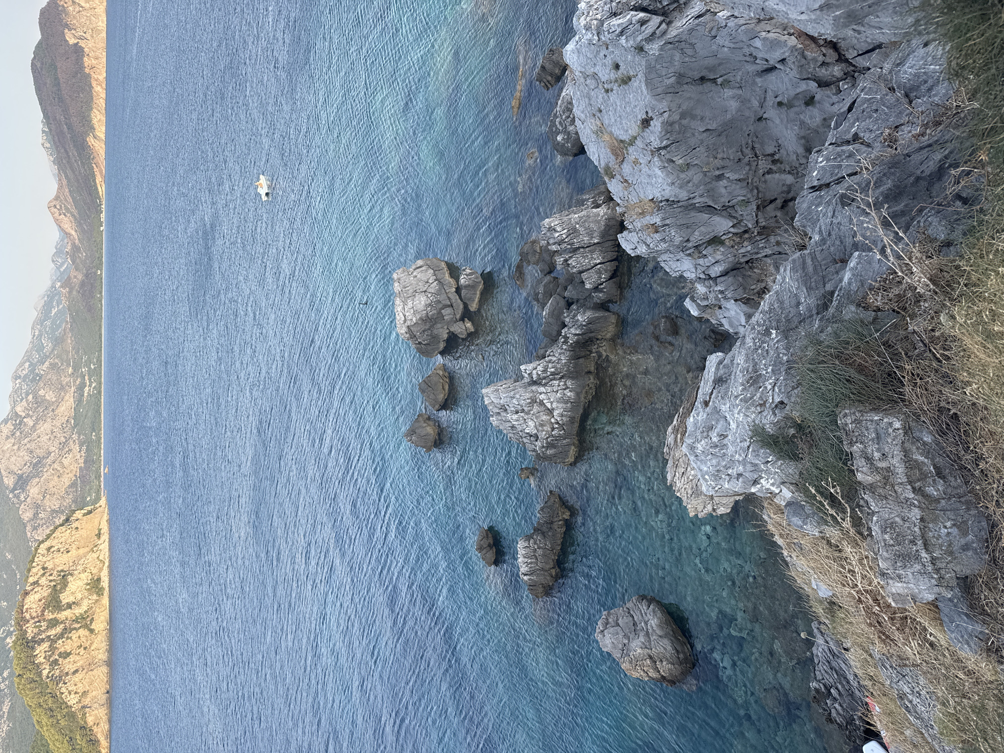 This is a high-angle, eye-level shot of a rocky coastline and the sea near Petrovac centar, Montenegro. In the foreground, large, grey rocks are covered in dry grass and vegetation. The water is a clear, bright turquoise, revealing submerged rocks and the seabed in shallower areas. Several larger rocks break the surface of the water. In the mid-ground, a small white boat with a person inside is visible on the water. In the background, a large, tree-covered hill juts out into the sea, and beyond that, a range of barren, mountainous terrain stretches across the horizon under a pale sky.