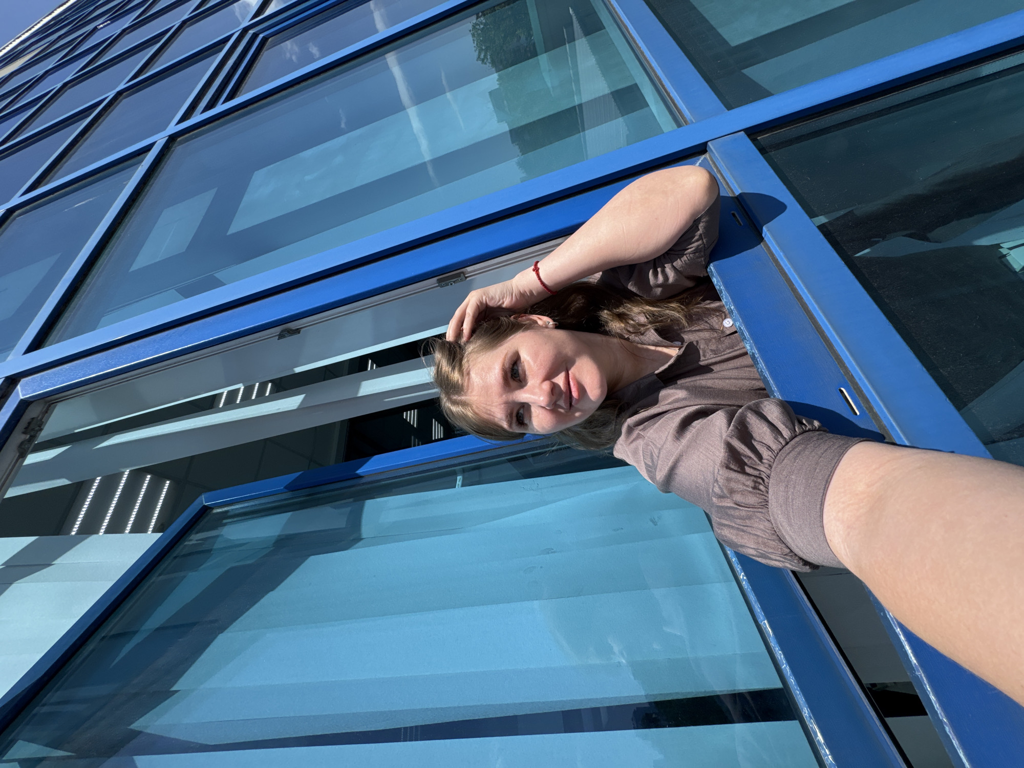 The image is taken from a low angle, looking up at a woman leaning out of an open window of a modern building. The building has a facade of large, blue-framed windows. The woman has fair skin and light brown hair, and she is wearing a long-sleeved, brown shirt. Her left arm is raised, and her hand is touching her hair. A red bracelet is visible on her left wrist. The windows reflect the sky and sunlight. Based on geotags, the location is Perm, Russia.