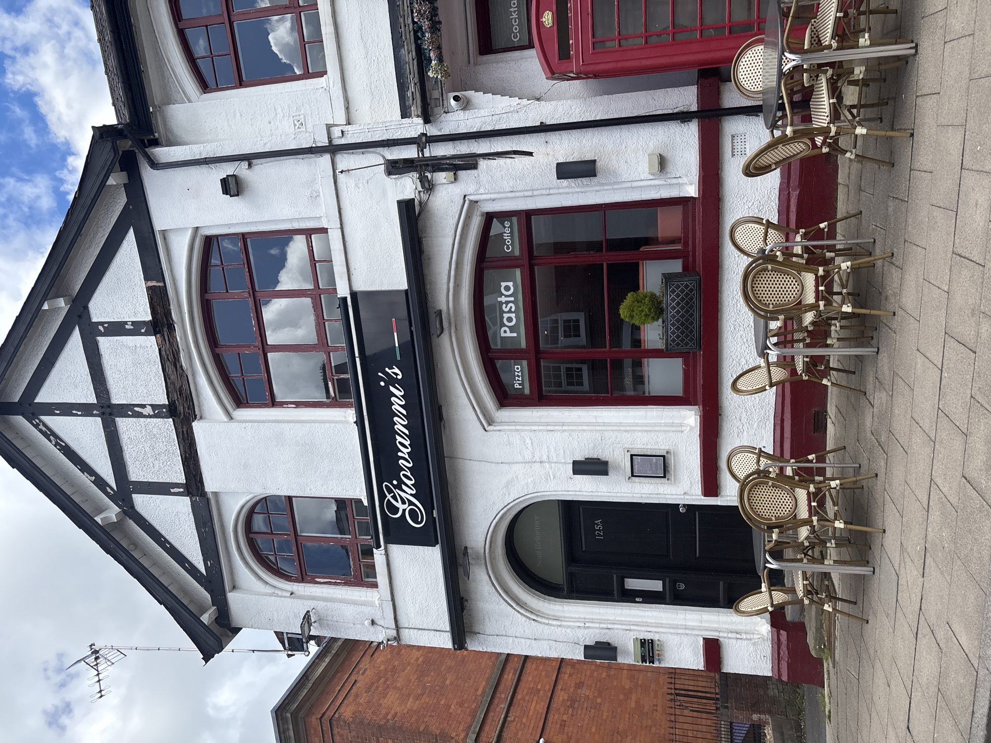 The image shows the exterior of Giovanni's restaurant in Knutsford, United Kingdom. The building is white with black timber framing on the left side, and red trim around the windows and door on the right. A black sign with "Giovanni's" in white script is mounted above the entrance. The arched entrance door is black and labeled "125A". Circular windows above the entrance have red frames and reflect the sky. Words "Pasta", "Pizza", and "Coffee" are printed on the windows, in the same manner as "Cocktails" in a window at the top right of the image. Several rattan chairs and small tables are arranged on the sidewalk in front of the building. A brick building is visible to the left and a wrought iron fence is visible to the right.