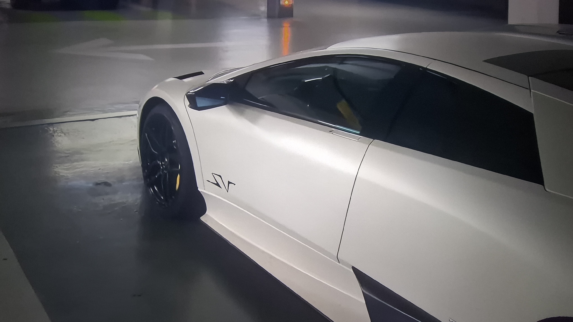 A partial view of a white Lamborghini Murciélago SV is captured in what appears to be an underground parking garage. The car is angled towards the upper right, revealing its side profile from the front wheel to the rear section of the roof. The black alloy wheel is partially visible on the left. The "SV" logo is visible on the side of the vehicle. The surrounding environment is a typical parking garage setting, with painted directional arrows on the floor and concrete structural elements. The location in the city of Unknown, Unknown.