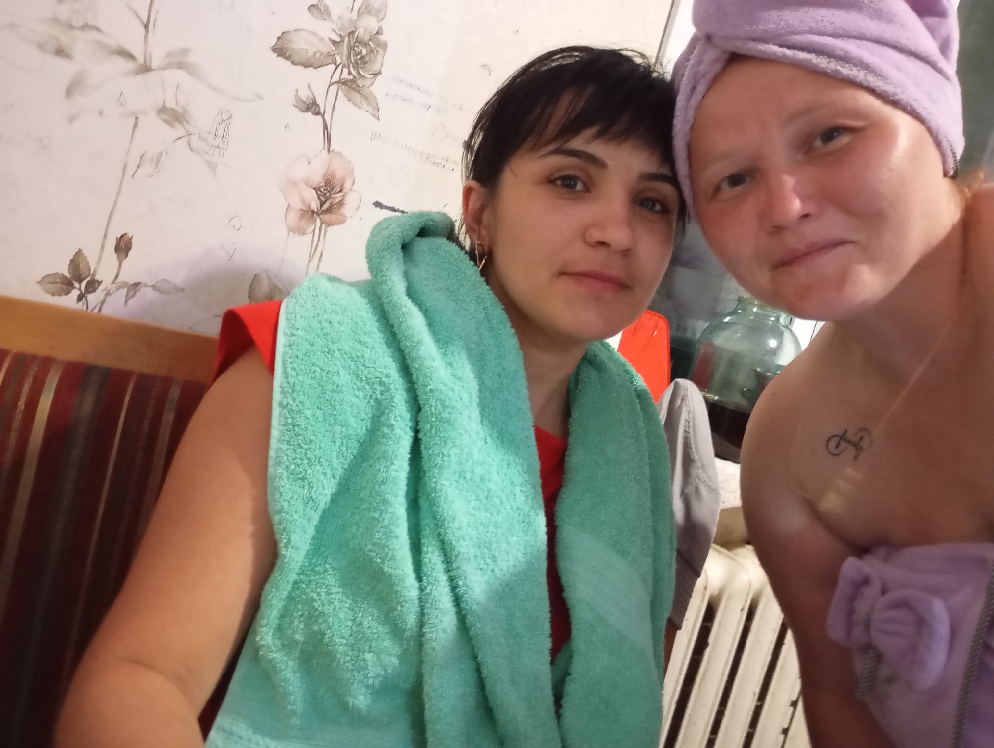 Two women are in a room. The woman on the left has dark hair and is wearing a red shirt with a teal towel draped over her. The woman on the right has blonde hair and is wearing a purple towel wrapped around her head. She also has a tattoo of a bicycle on her shoulder. A large glass jug is visible behind the woman on the right. The room has floral wallpaper and a striped upholstered bench. The scene appears to be indoors, likely in a home. The exact location in the city of Ryabki, Russia is not specified in the image.