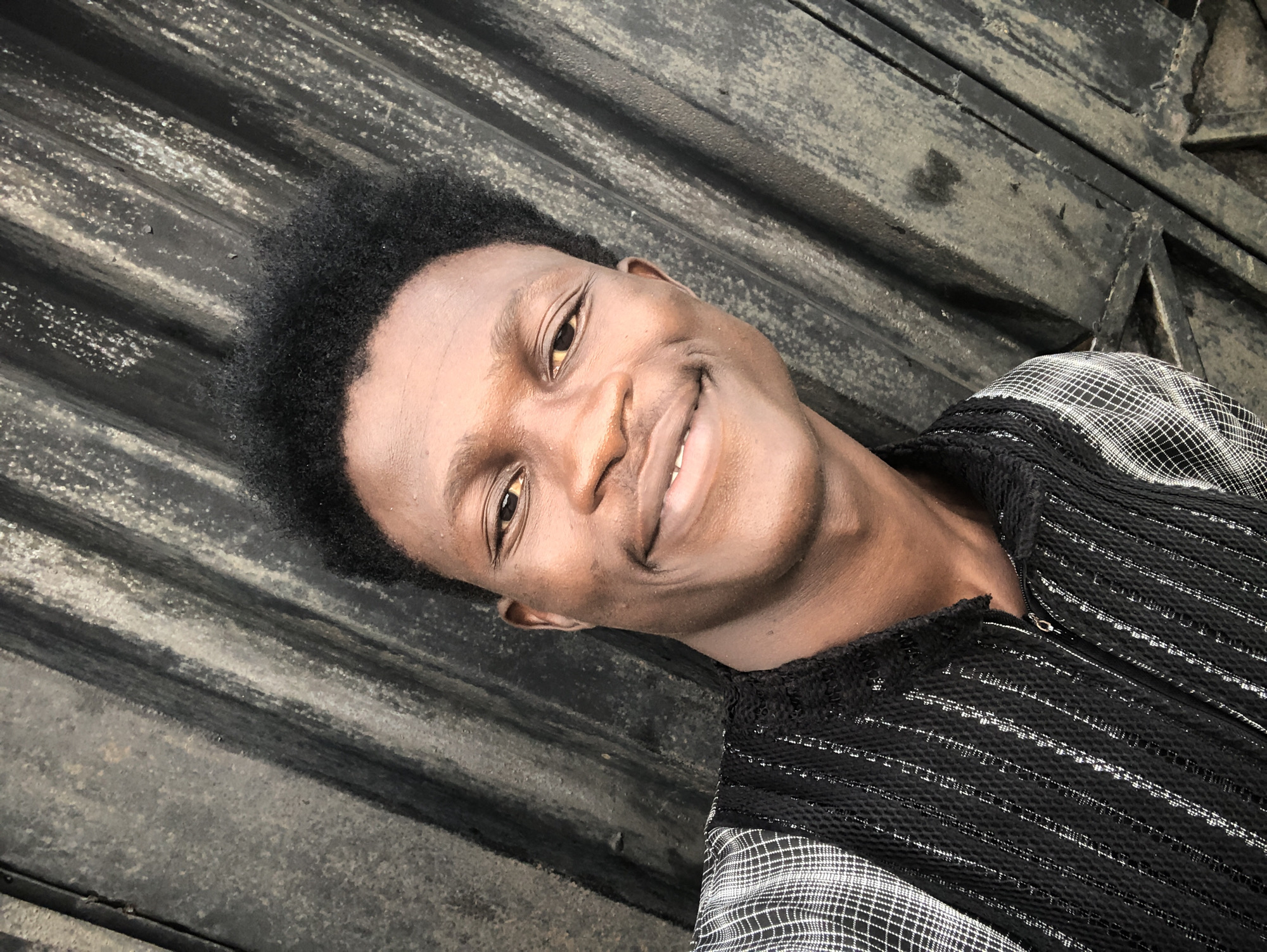 A medium shot captures a young Black man with dark, curly hair, smiling broadly at the camera. He is wearing a black shirt with a checkered pattern on the shoulders and sleeves, and a textured, ribbed fabric with vertical white stripes on the chest. The background is a dark, corrugated metal surface, possibly a gate or wall, with a distressed and weathered appearance. The lighting suggests an outdoor setting during the day. The overall impression is a casual, candid portrait.
