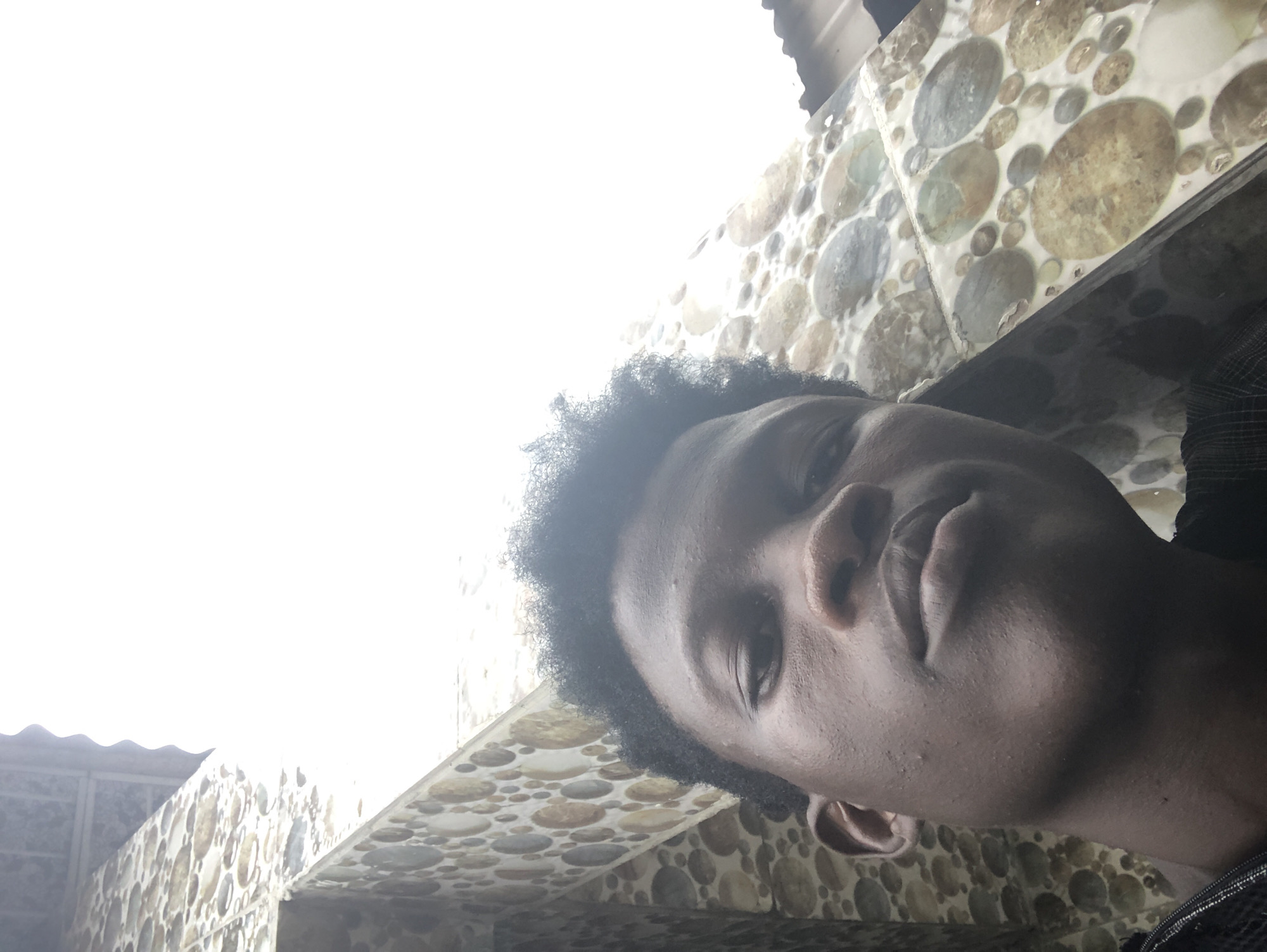 A young Black male with dark skin and short, dark, curly hair takes a close-up selfie from a low angle. He is looking directly at the camera with a neutral expression. His face is well-lit, showing some texture and a few blemishes on his skin. He is wearing a dark, collared garment, possibly a shirt or jacket, with a subtle striped pattern visible.
The background consists of a tiled wall with a circular pattern of stones in various shades of brown, gray, and white. The wall appears to be part of an exterior building. Above the wall, a corrugated metal roof is visible, and beyond that, a brightly overexposed sky dominates the upper portion of the image. The lighting suggests it is daytime, possibly very sunny given the blown-out sky. The setting is an outdoor environment, possibly a residential or urban area. There is no specific identifiable landmark to confirm the location within Ado, Nigeria, but the overall architecture and material choices are consistent with common construction styles in the region.