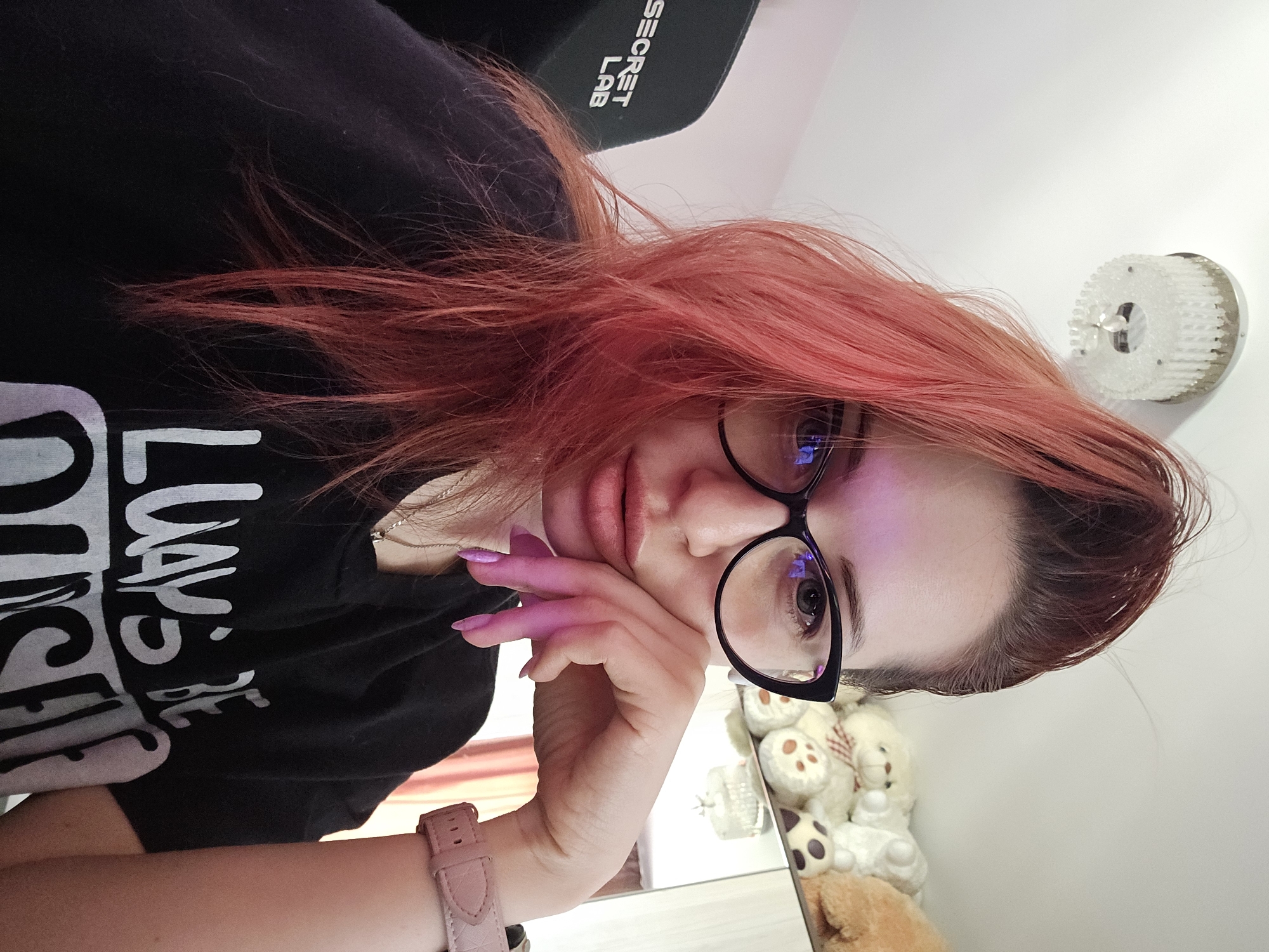 A woman with red hair and glasses is looking at the camera while resting her chin on her hand. She is wearing a black t-shirt with white text on it. A pink watch is visible on her wrist. The background includes a ceiling lamp, stuffed animals, and a chair with "SECRET LAB" printed on it. The scene is indoors.
