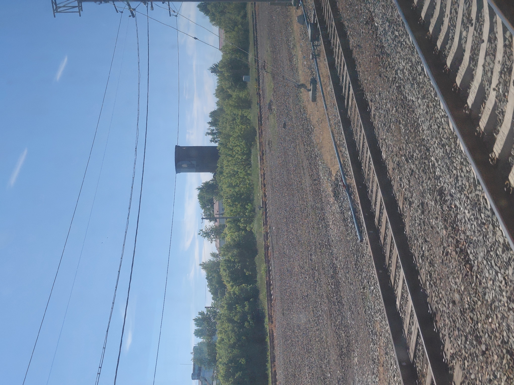 This image depicts a railway scene with several tracks running parallel to each other. The foreground is dominated by the ballast of the railway, with wooden sleepers visible beneath the metal rails. In the middle ground, there is a dense line of green trees, behind which are some buildings, including what appears to be a tall, cylindrical structure resembling a water tower. The sky is a clear blue with a few faint wispy clouds. Overhead, power lines are strung across the frame. The scene suggests an industrial or semi-rural area, likely on the outskirts of a settlement. Based on the provided context, this scene is located in the city of Chulym, Russia. There are no people or animals visible in the image.