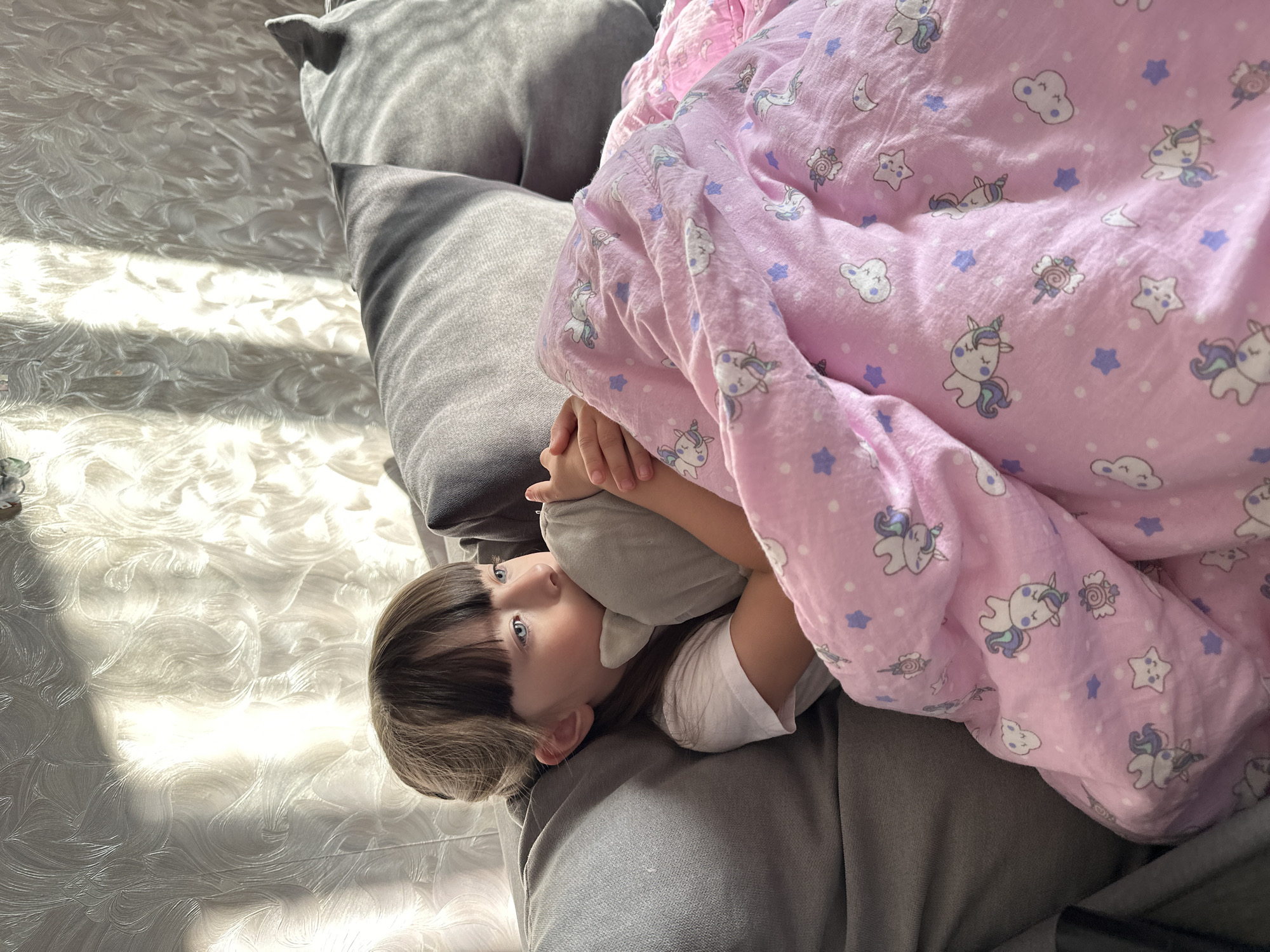 A young child with dark bangs and shoulder-length brown hair is lying on a gray couch with several pillows. The child is mostly covered by a pink blanket decorated with unicorns, stars, and clouds. The child is holding a gray stuffed animal close to her chest and looking upwards to the left. Sunlight illuminates a patterned floor next to the couch. The scene appears to be indoors. The location of Chaikovsky, Russia is not discernable from the image.