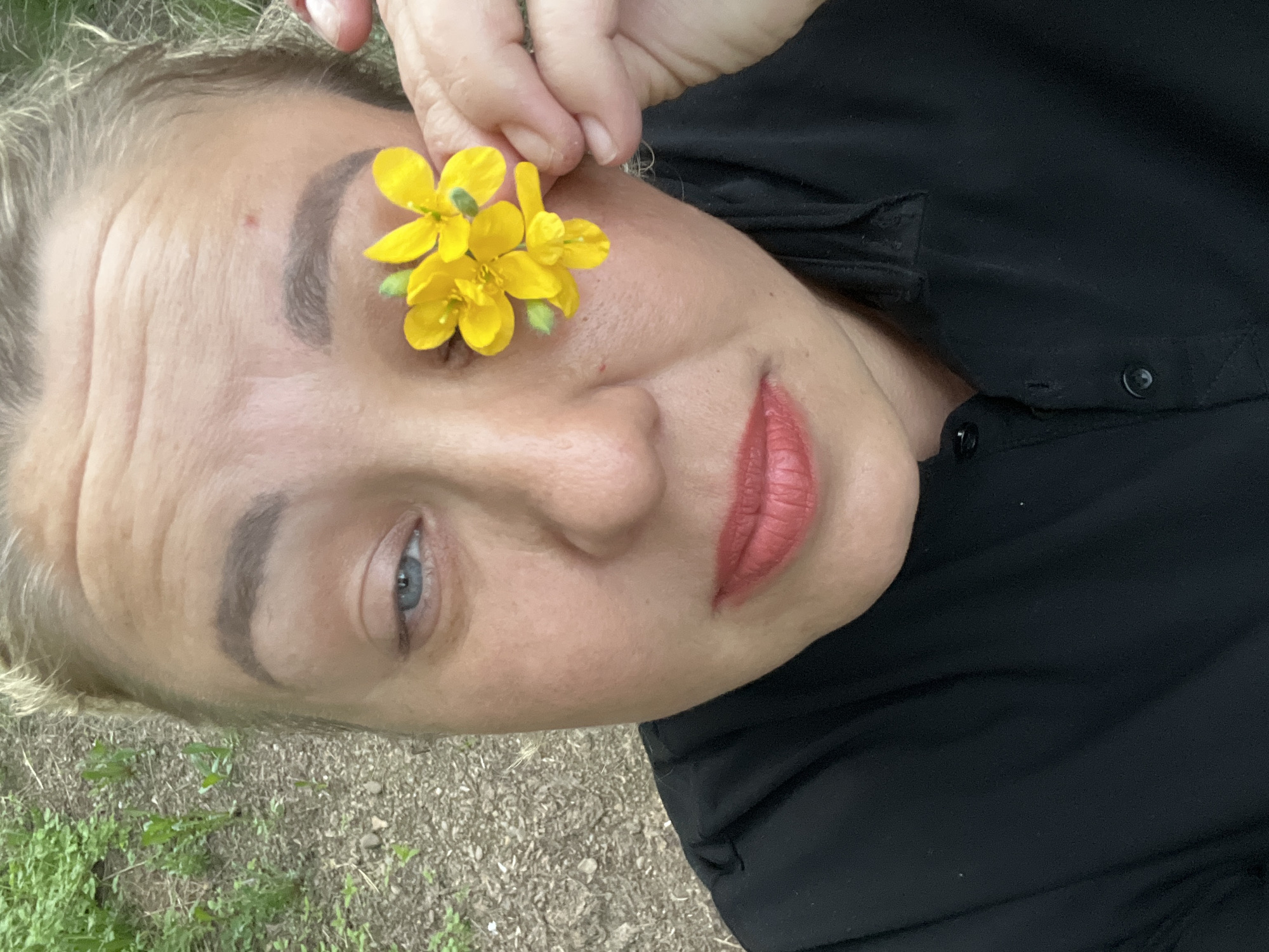 The image shows a person with fair skin lying on the ground, likely outdoors, given the presence of green vegetation and what appears to be gravel or dirt in the background. The person is wearing a black, button-down shirt. The person is holding a small cluster of bright yellow flowers near their left eye. Their hair appears to be light-colored and partially visible. The lips are colored with what appears to be a reddish-coral lipstick. The eye that is visible appears to be blue or gray. It is not possible to confirm the location as Chaikovsky, Russia, from the given image.