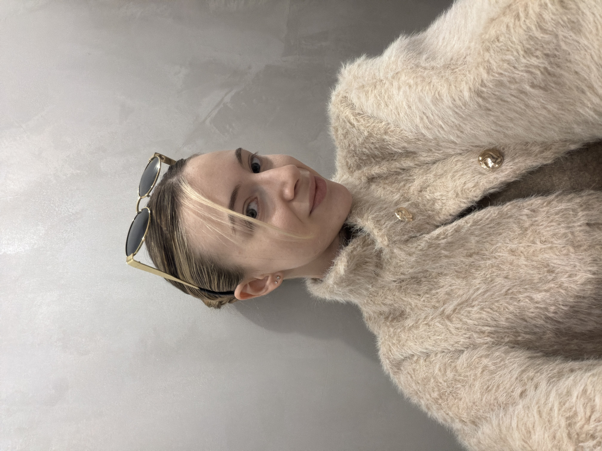 A blonde woman with sunglasses perched atop her head, wears a fuzzy beige coat with gold buttons. She is indoors, posed against a neutral gray wall. The location in the city of Plovdiv, Bulgaria is not identifiable from the image.