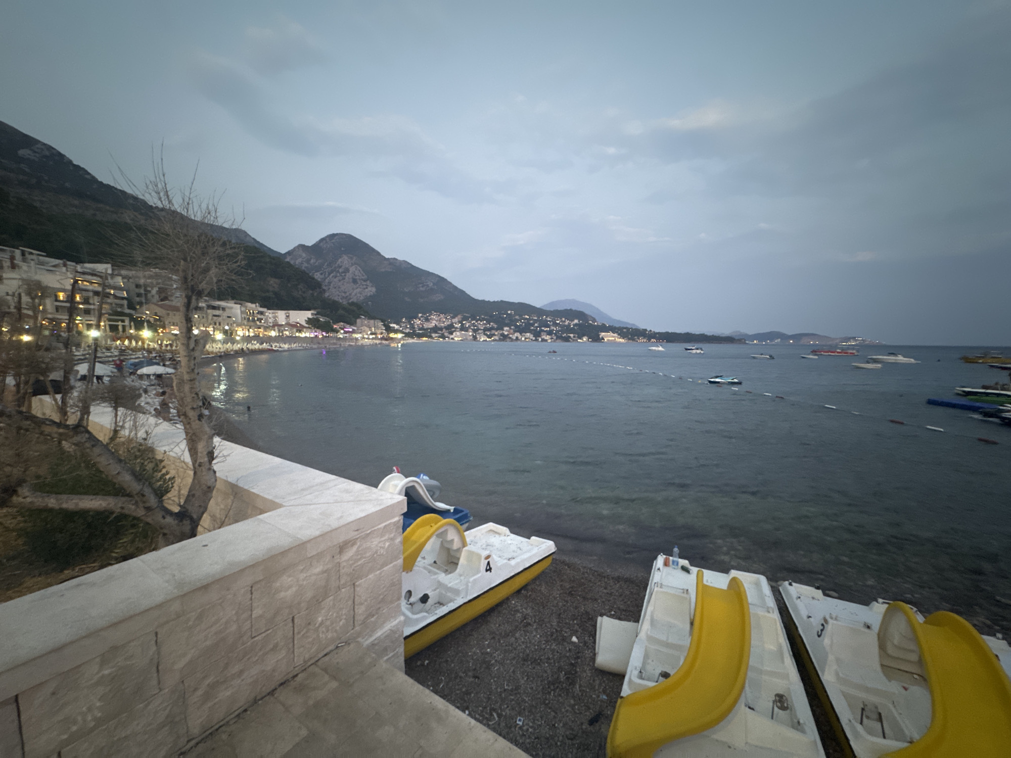 The image shows a dusk scene at a beach in Sutomore, Montenegro. The foreground features a stone wall and a bare-branched tree. To the right, two pedal boats with yellow slides are pulled up onto the pebble beach. In the middle ground, the calm sea is dotted with several boats, including a jet ski. The background reveals a long stretch of beach lined with buildings and hotels, backed by dark, mountainous terrain. The sky is overcast with a gradient of gray and lighter tones. The overall impression is of a coastal resort town at the end of the day, with an atmosphere of quietude before nightfall.