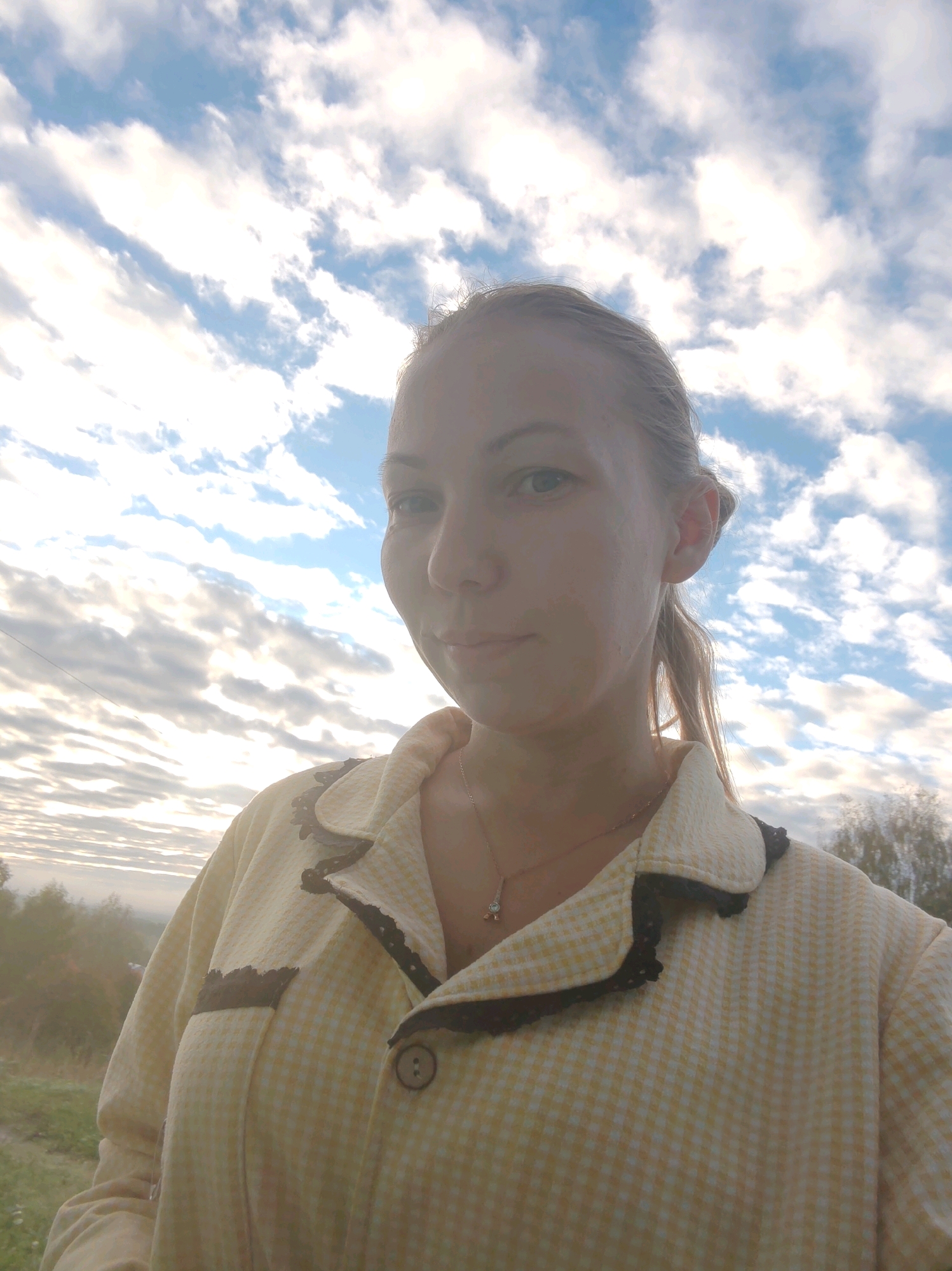 A close-up selfie shows a young woman outdoors against a cloudy sky. She is wearing a light yellow, checkered collared shirt with dark trim and a small button closure. A delicate necklace with a pendant is visible on her chest. The background features a sky with scattered white clouds and hints of trees and landscape at the bottom edge. The lighting suggests it is either early morning or late afternoon. The overall setting appears to be rural, with no visible city structures or landmarks to suggest it is specifically in Чухлэм, Russia.