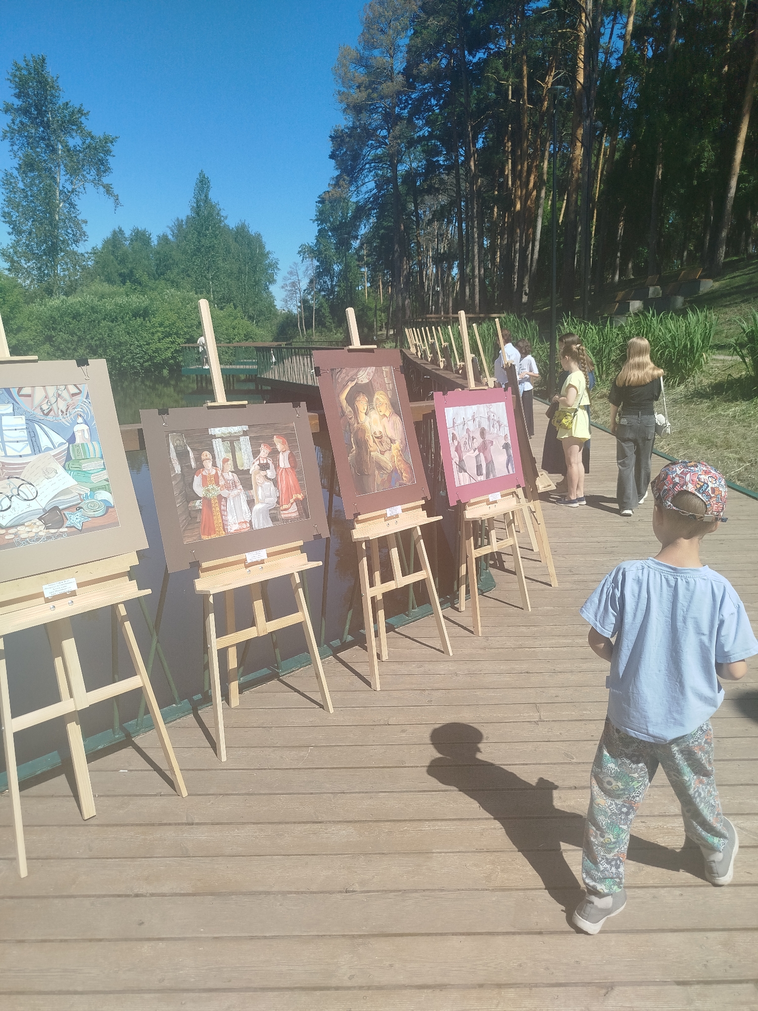 In Zavodoukovsk, Russia, a temporary outdoor art exhibition is taking place on a wooden boardwalk adjacent to a body of water. Several paintings are displayed on wooden easels, featuring various subjects, including still life, historical scenes with figures in traditional attire, and what appears to be a depiction of children playing.
A young boy, wearing a light blue t-shirt and patterned trousers, stands with his back to the camera, looking at the artwork. Behind him, a group of people, including women and possibly men, are also observing the exhibition. The environment is characterized by lush green trees and clear blue skies, suggesting a pleasant day. The boardwalk is made of weathered wooden planks, and a green metal railing runs along the edge of the water. The overall scene conveys a community event focused on art in a natural, scenic setting.