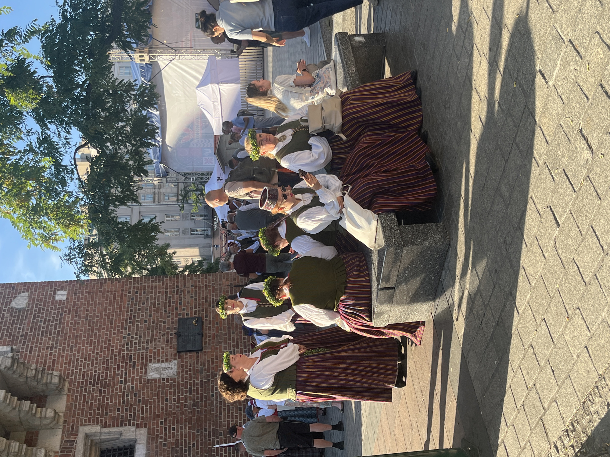 The image captures a sunlit scene in Krakow, Poland, likely near the Wawel Castle due to the prominent brick structure with carved stone elements. Several people are present, many dressed in traditional folk costumes featuring floral wreaths, white blouses, and long, striped skirts in shades of red and purple.
In the foreground, three women in folk attire are seated on a stone bench. The woman on the left, wearing a conical hat and sunglasses, is looking at her phone. The woman in the middle is also seated, holding her phone, and wearing a wreath. The third woman is seated to their right, facing away from the camera, also wearing a wreath and folk attire.
To the left of the seated women, two other women in similar folk costumes stand talking. One is wearing a wreath and a white blouse with a dark vest and a striped skirt. The other, also in folk attire, is turned away from the camera.
Further back, a man in a green shirt and black shorts stands holding what appears to be a spear or flagpole. Other people in casual clothing are visible in the background, along with white tents and scaffolding, suggesting an outdoor event or festival. The ground is paved with stone slabs, and shadows from trees and structures are cast across the scene. The sky is clear and blue.