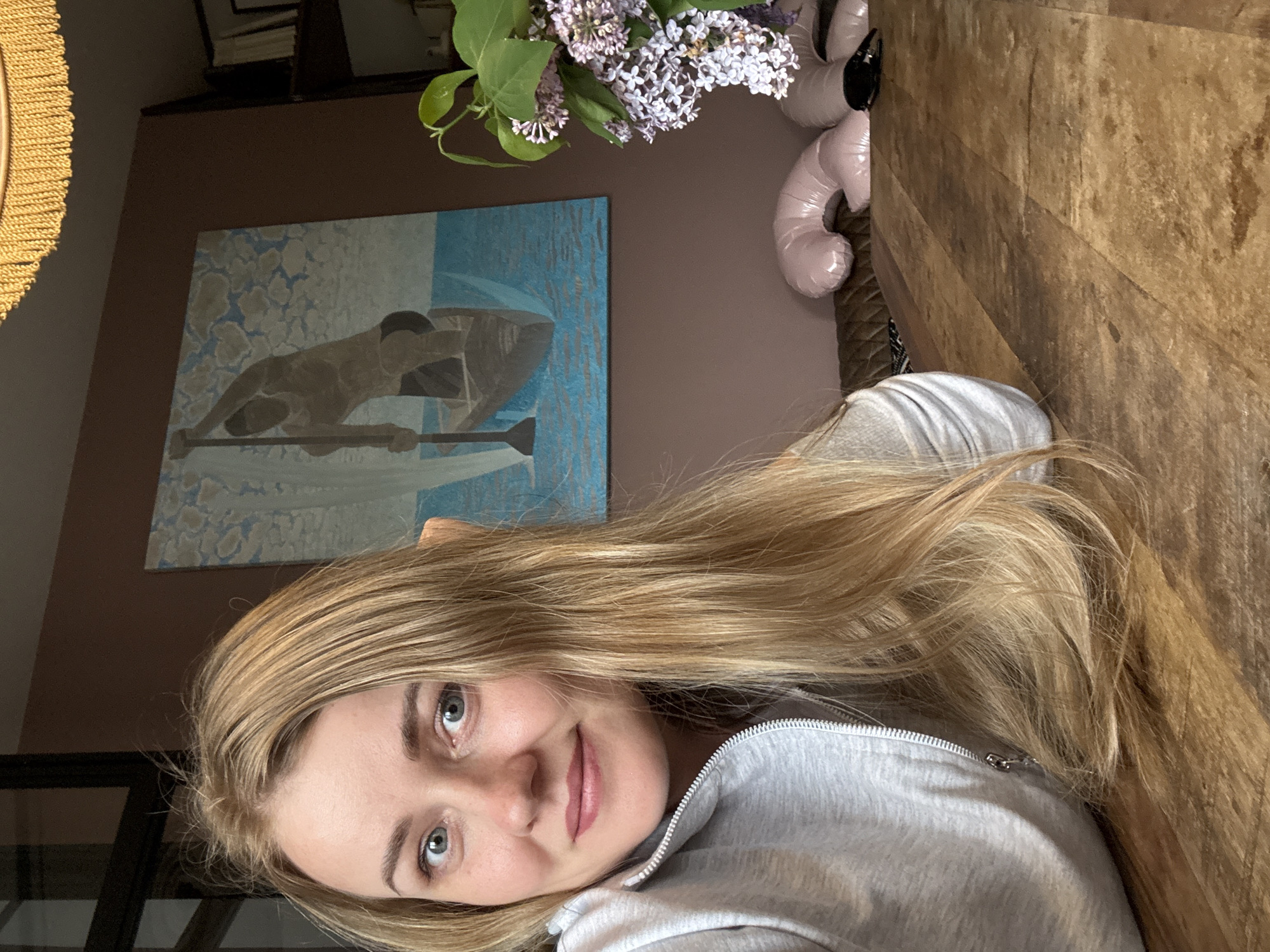 A fair-skinned woman with long, blonde hair and light-colored eyes is centered in the image, wearing a gray zip-up sweatshirt. She faces the camera, slightly tilted. Behind her, a light brown wall is visible with an artwork depicting a figure in a boat. A bouquet of lilac flowers is placed on a table, and a pink, inflatable object is partially visible to the right. The table is made of light brown wood. There is not enough information to determine the location in the city of Saint Petersburg, Russia.