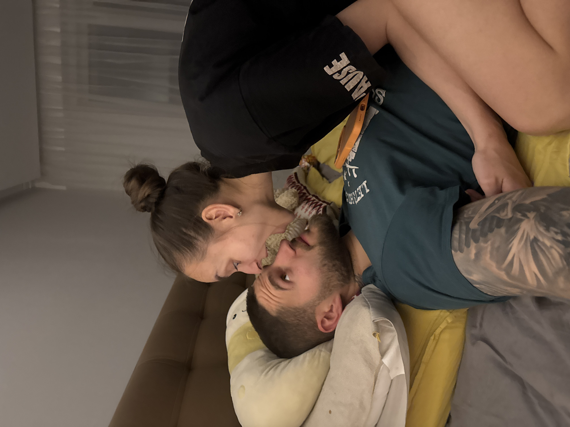 A woman and a man are lying in bed, presumably in a private residence. The location is unknown but suggested to be Sofia, Bulgaria. The woman, with her hair in a bun, is leaning over the man and appears to be kissing him on the face. She is wearing a black t-shirt. The man is lying on a pillow, wrapped in bedding. He has a beard and tattoos on his arm. The bed is covered in sheets and a yellow blanket. There is a cell phone on the bed and various pillows. The wall is painted a light neutral color.